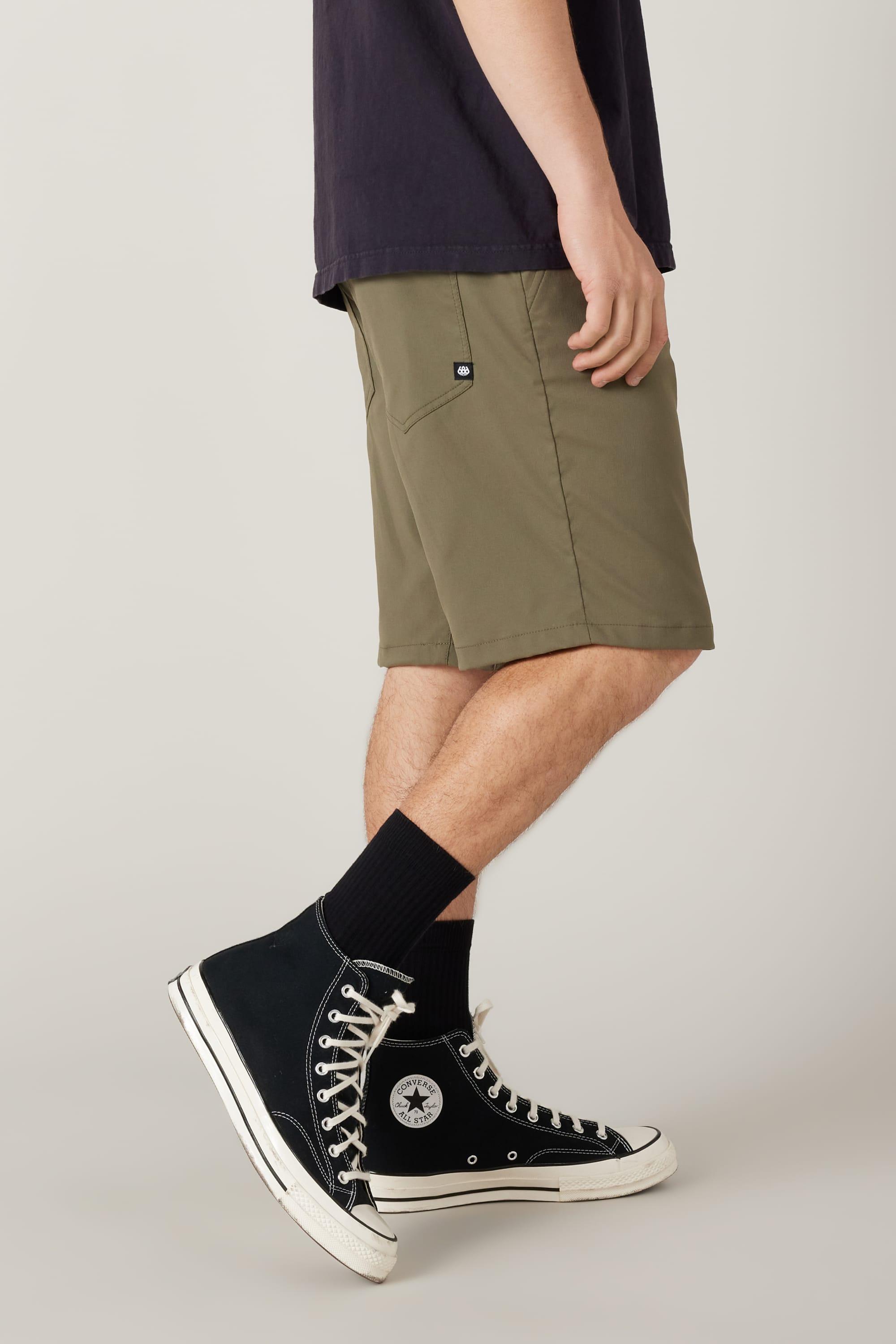 A person stands sideways against a neutral background wearing 686 Men's Everywhere® Hybrid Short - Relaxed Fit in olive green, paired with a black t-shirt, black crew socks, and black high-top Converse sneakers.