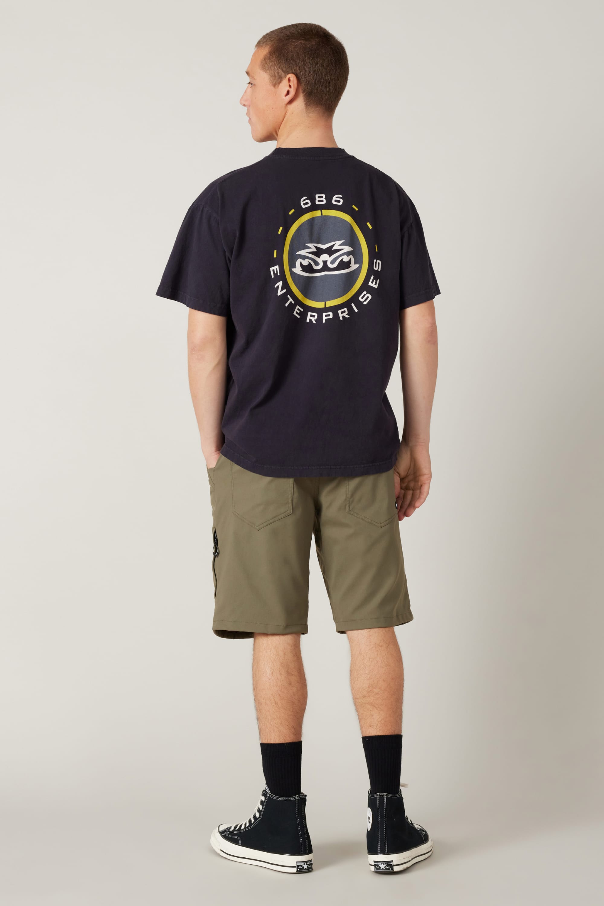 A person stands facing away, wearing a black t-shirt with a yellow and white 686 Enterprises logo, 686 Men's Everywhere® Hybrid Short - Relaxed Fit in olive green, black socks, and black Converse sneakers against a plain background.