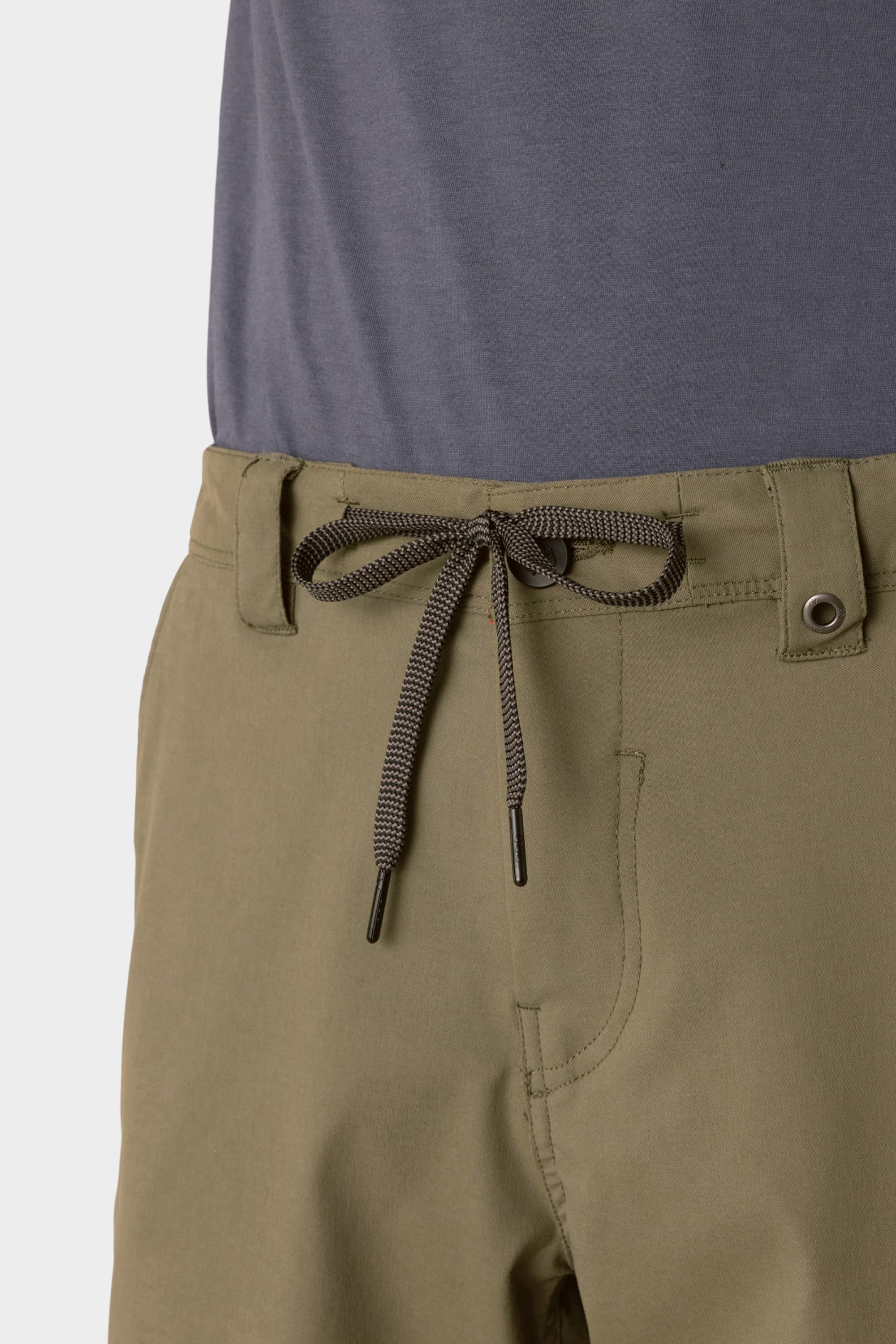 A close-up shows a person wearing 686 Men's Everywhere® Hybrid Short - Relaxed Fit in olive green with a black-and-white drawstring, paired with a tucked-in grayish-blue shirt.