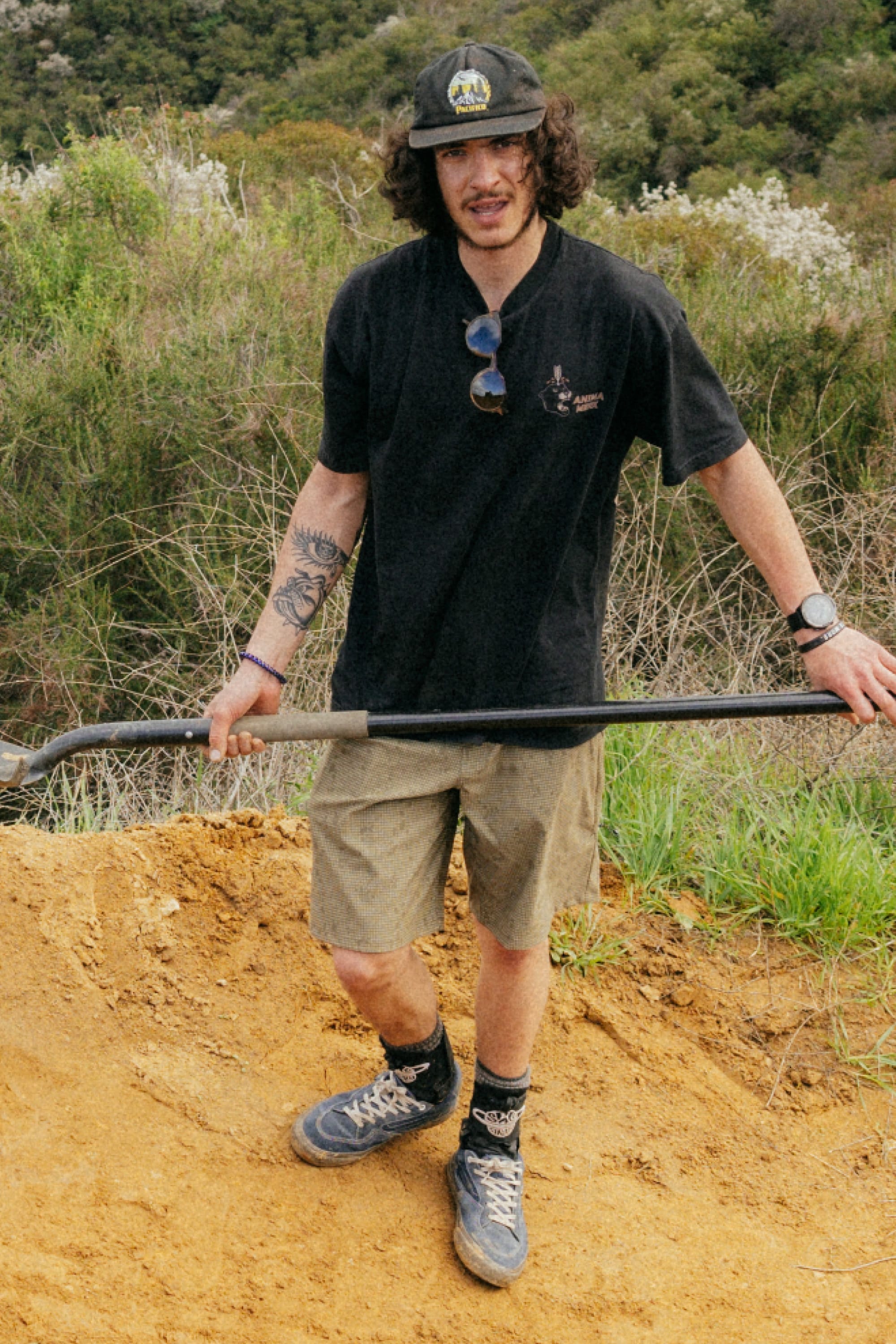 A person wearing a black hat, black t-shirt, 686 Men's Everywhere® Hybrid Short - Relaxed Fit, and sneakers stands on a sandy path with a long-handled tool, surrounded by greenery and bushes.