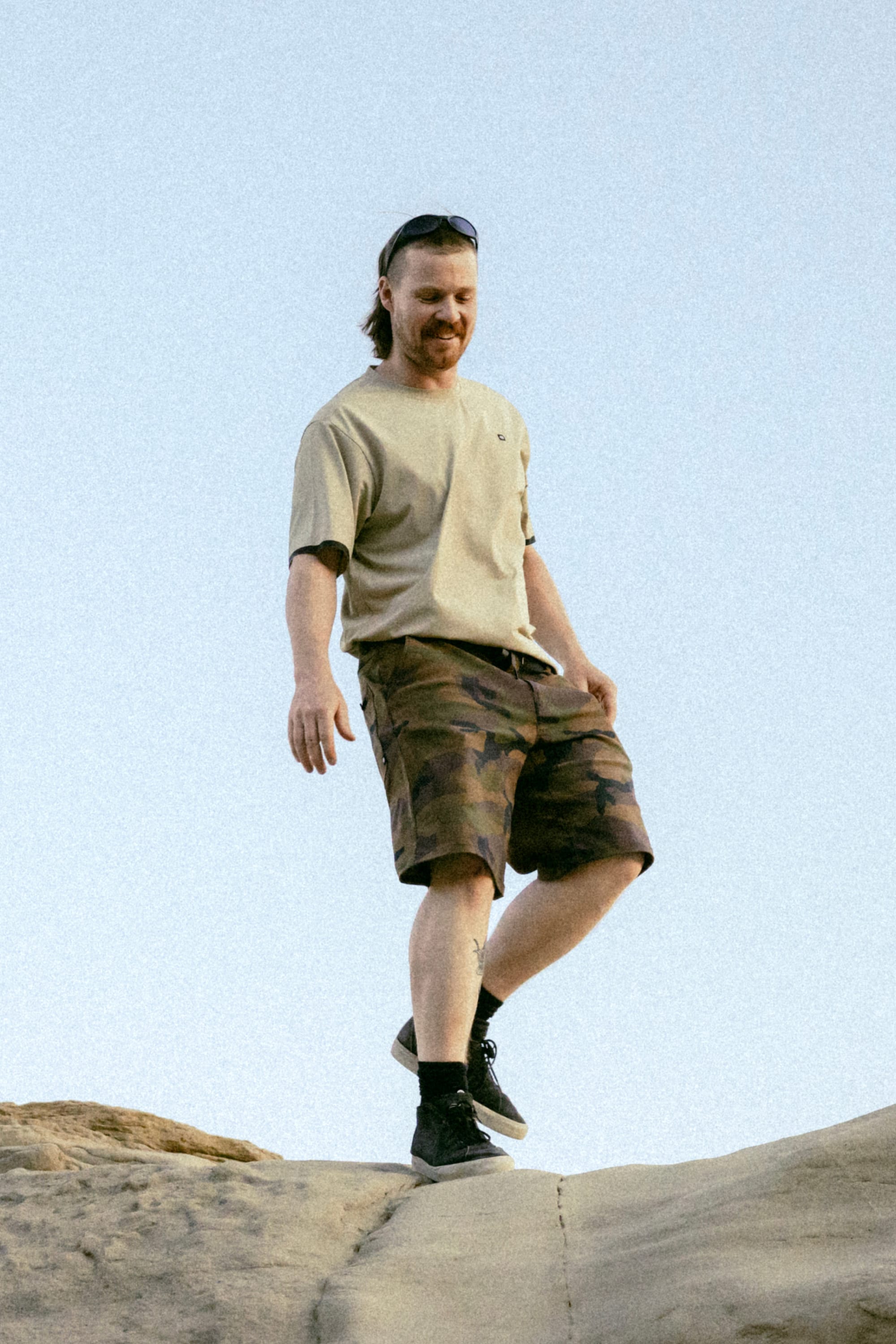 A man, smiling and looking down, stands on a rock under a clear blue sky outdoors wearing beige shorts by 686 Men's Everywhere® Hybrid Short - Relaxed Fit, a beige t-shirt, and black shoes.