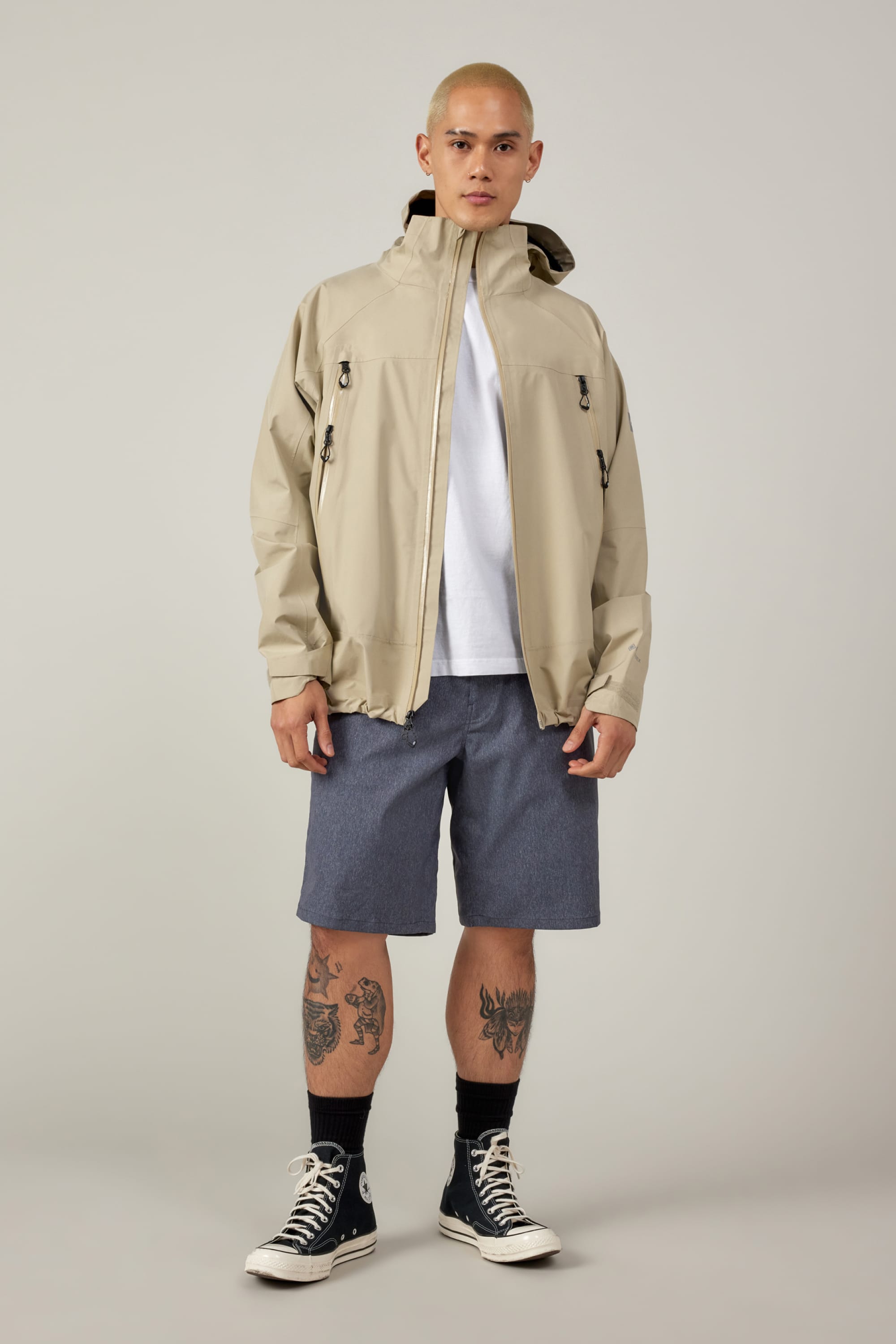 A person with a shaved head wears a beige hooded jacket, white t-shirt, black socks, black Converse, and 686 Men's Everywhere® Hybrid Short - Relaxed Fit by 686. Tattoos are visible on both legs. They stand against a plain light background.