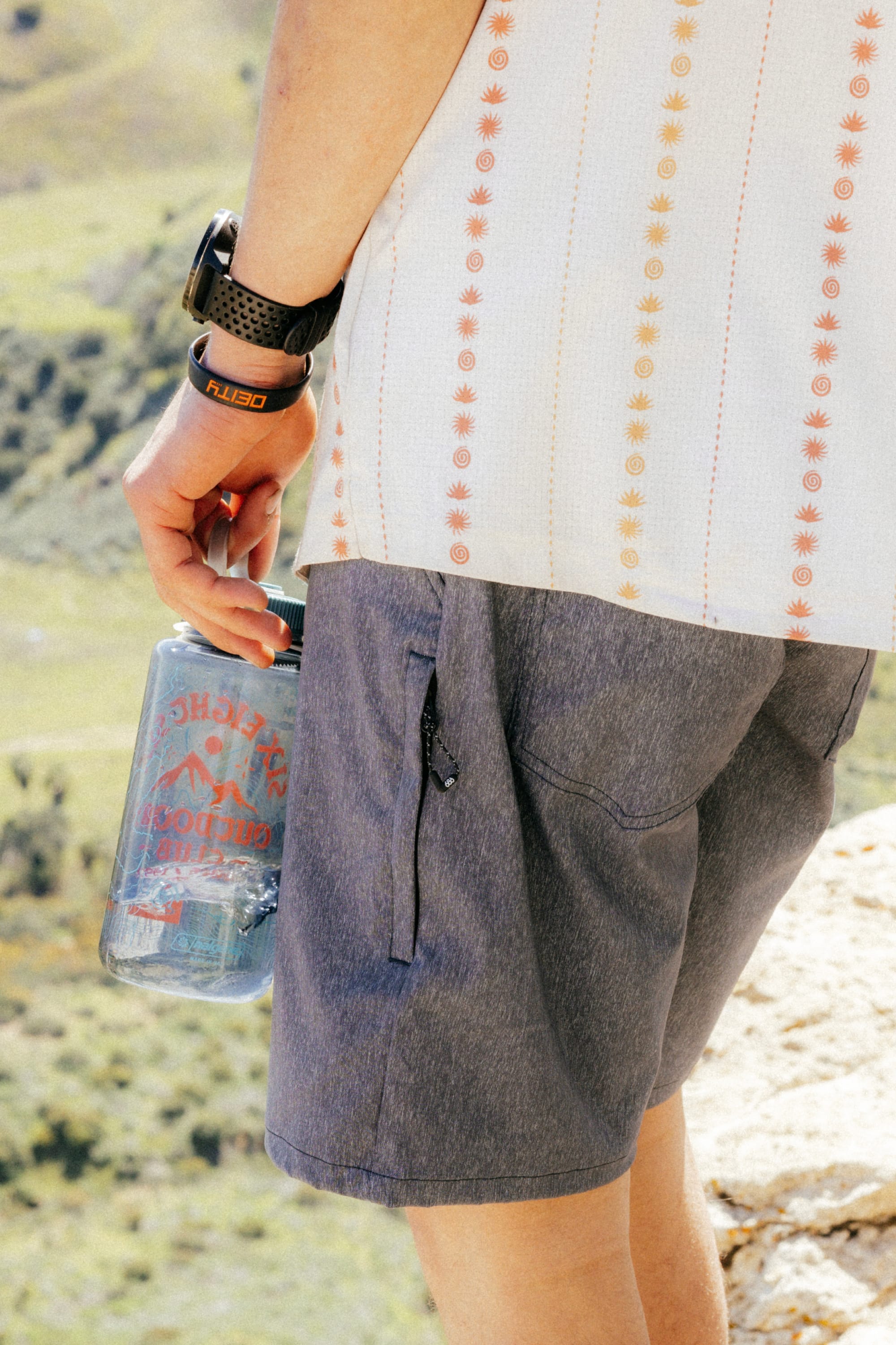 A person stands outdoors in 686 Men's Everywhere® Hybrid Short - Relaxed Fit, holding a large clear water bottle with a smartwatch on their wrist and a scenic, sunlit landscape in the background.