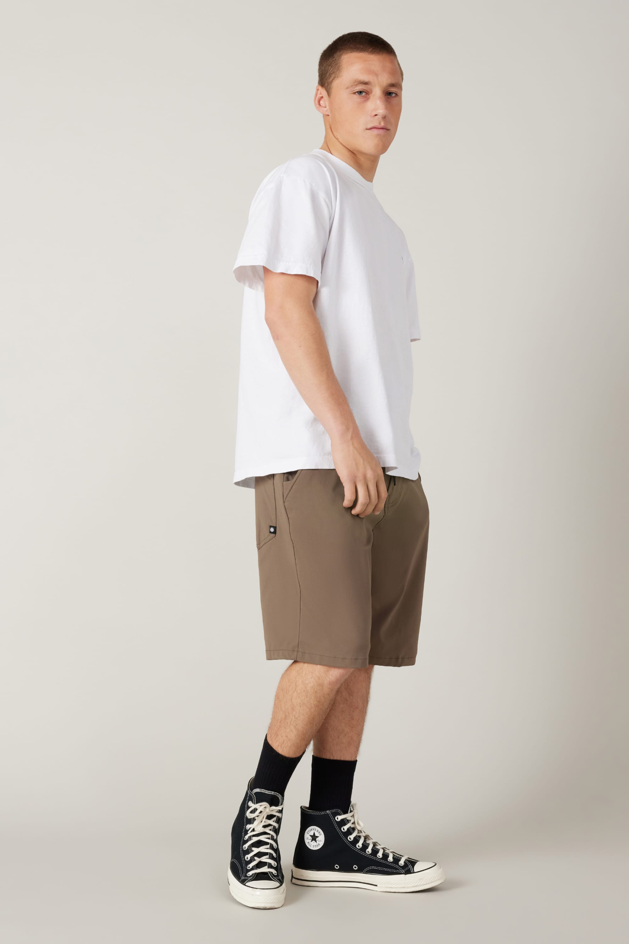A young man stands sideways, looking at the camera. He wears a plain white t-shirt, 686 Men's Everywhere® Hybrid Short - Relaxed Fit by 686, black crew socks, and black high-top Converse sneakers against a neutral background.