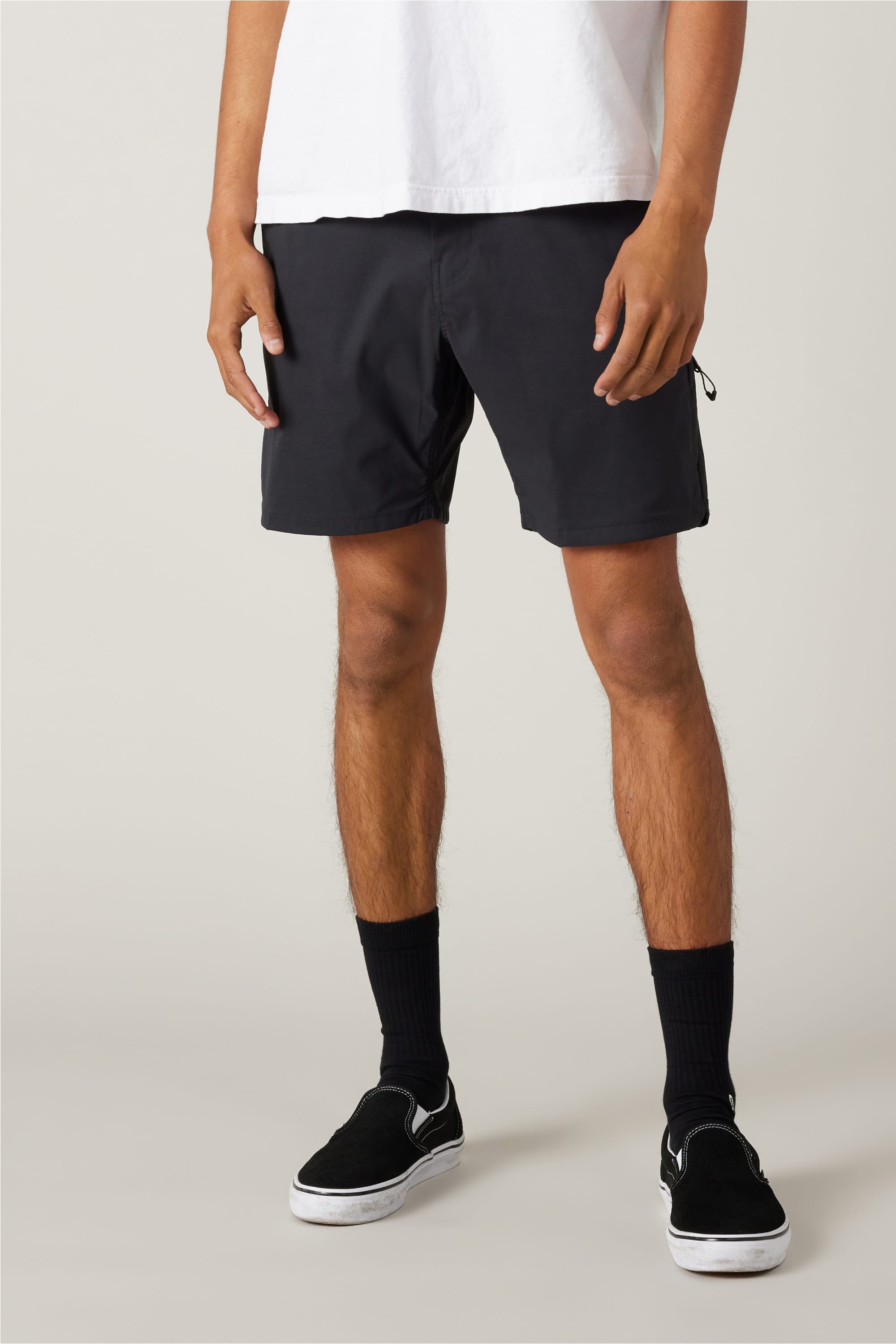 A person wears a white t-shirt, 686 Men's Everywhere® Featherlight Chino Short by 686 in black, black crew socks, and black slip-on sneakers against a plain light background. Their head is not visible in the image.