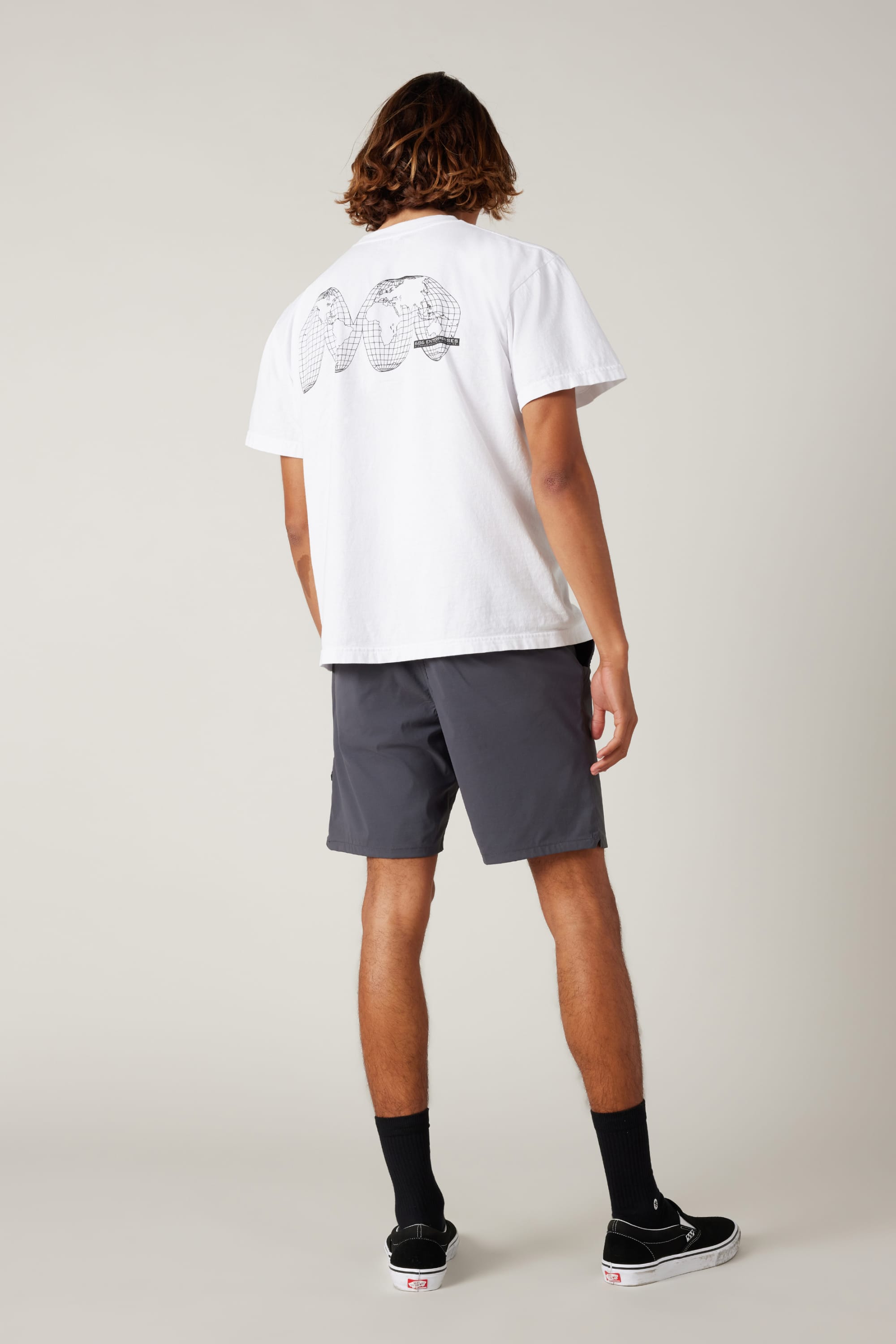 A person with shoulder-length hair wears a white graphic tee, 686 Men's Everywhere® Featherlight Chino Shorts by 686, black socks, and black sneakers. Facing away, the shirt’s back shows spherical designs on a plain light background.