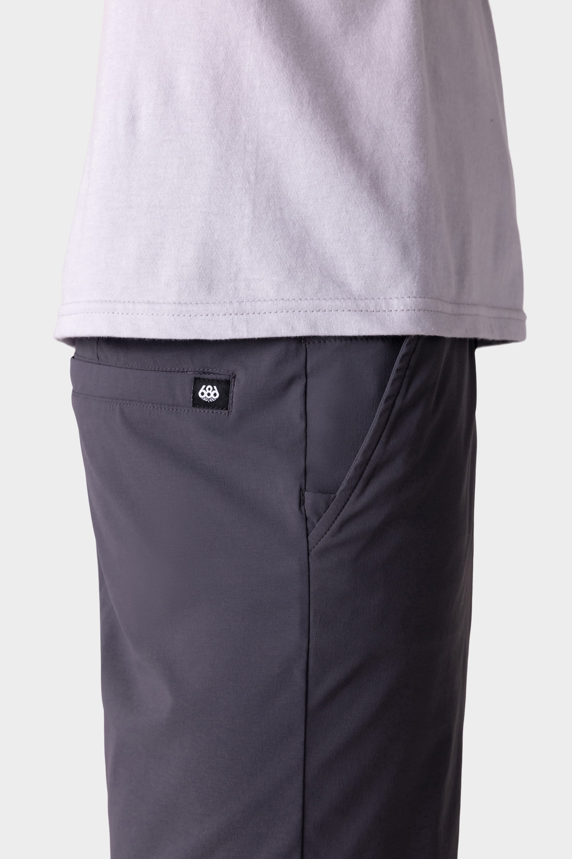 A side view shows a person wearing a light gray T-shirt and 686 Men's Everywhere® Featherlight Chino Shorts in dark gray, with a visible black and white 686 brand tag above the pocket. The background is plain and light-colored.