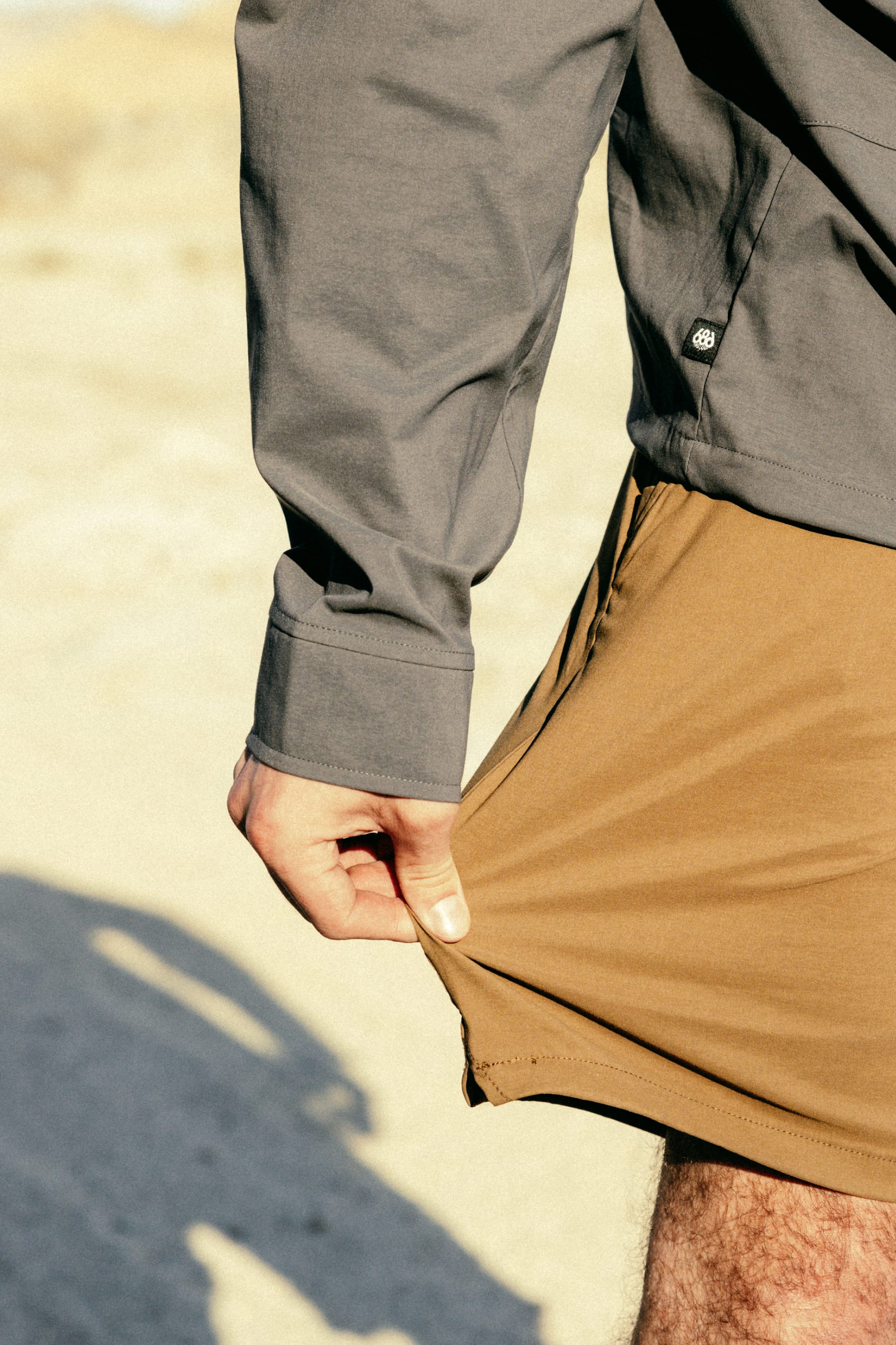 A person wearing a gray long-sleeve shirt and 686 Men's Everywhere® Featherlight Chino Short stands outdoors on sandy ground, gently pulling the hem to show the comfort and breathability of these lightweight shorts from 686.