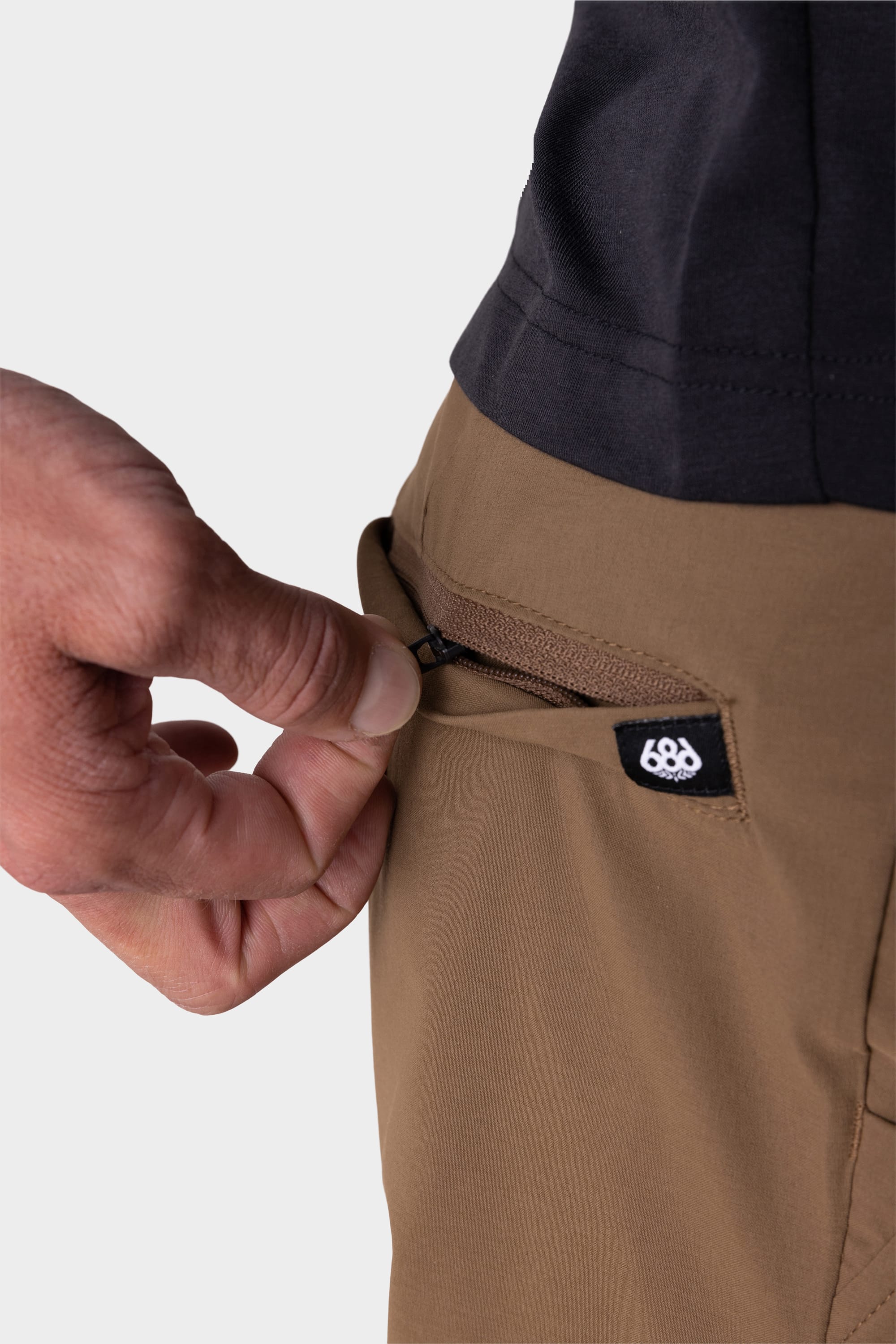 A close-up of a hand opening the zippered pocket of the 686 Men's Everywhere® Featherlight Chino Short in brown, featuring a small black and white 686 logo tag by the zipper. The person is wearing a black shirt.