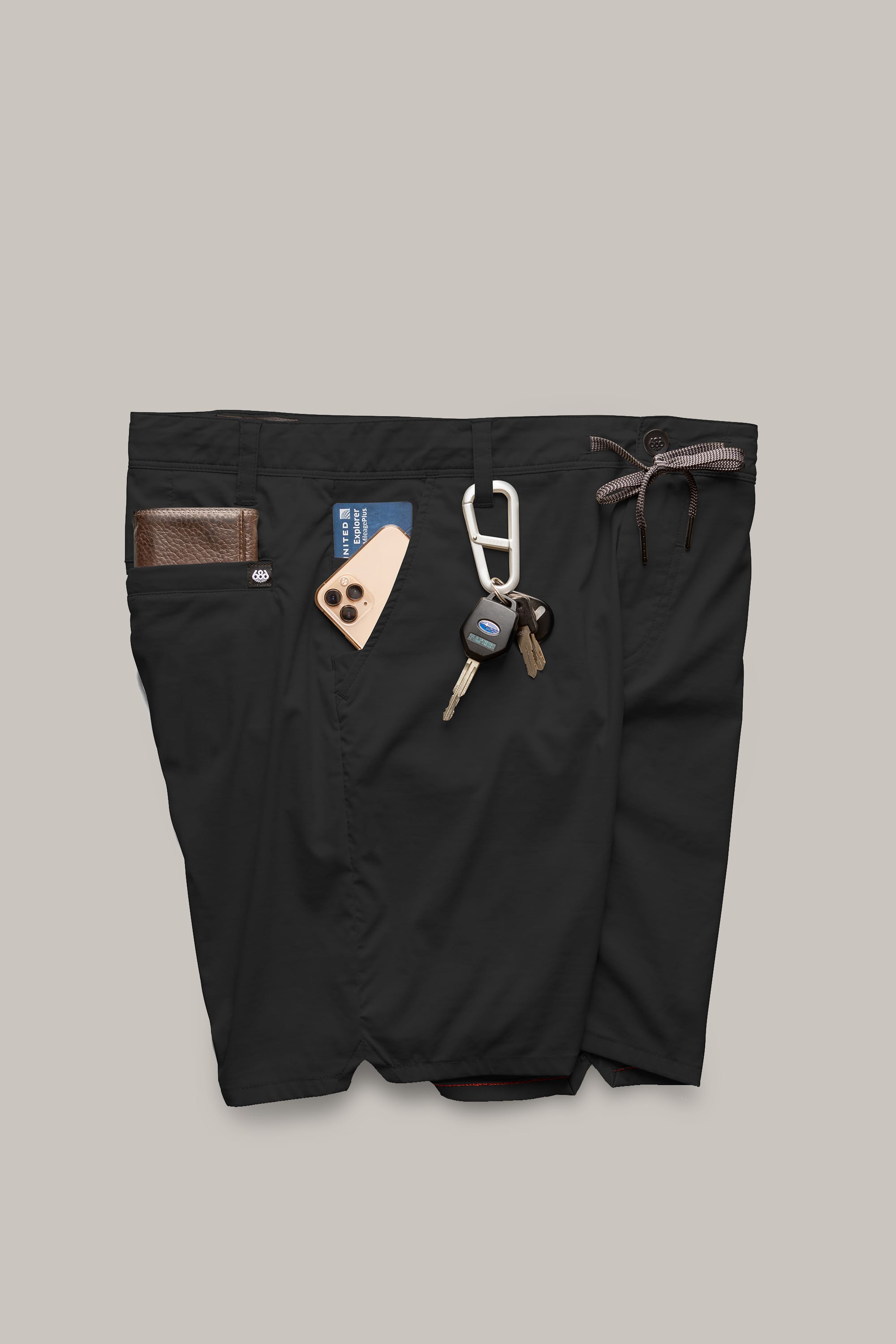 A pair of 686 Men's Everywhere® Featherlight Chino Shorts in black with a brown wallet in the left pocket, a phone and blue card in the right pocket, and keys on a carabiner hanging from the waistband.