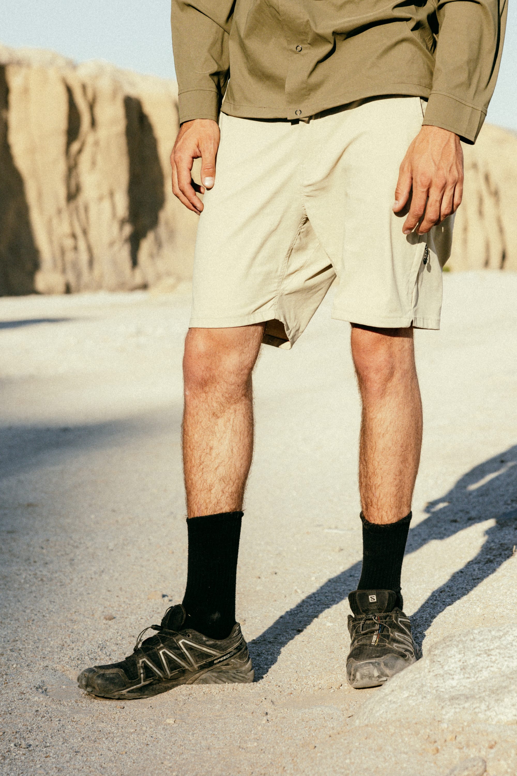 A person stands on a sunlit rocky terrain with cliffs in the background, wearing 686 Men's Everywhere® Featherlight Chino Short, a green shirt, black socks, and black trail shoes. The photo is cropped above the shoulders.
