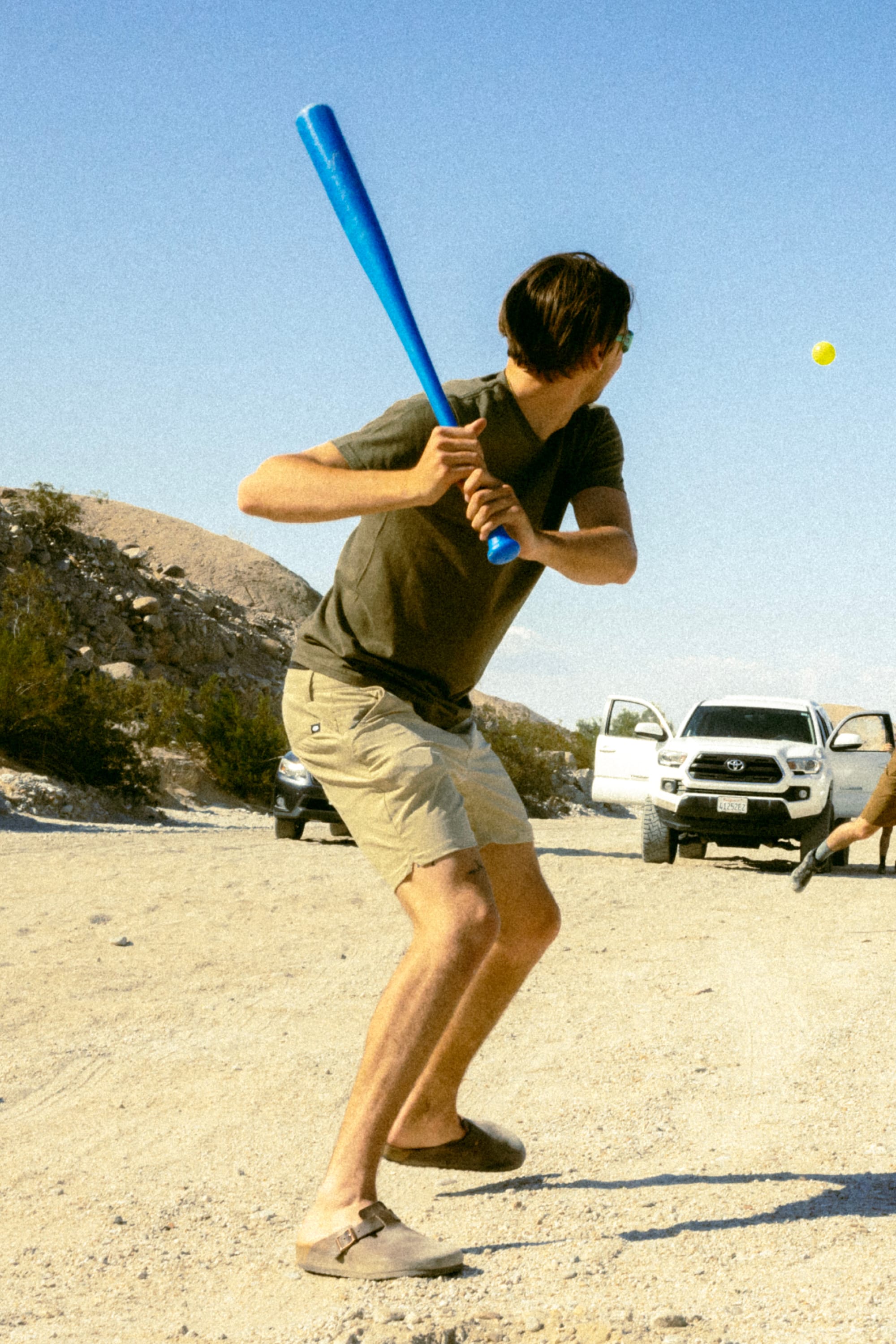 Wearing 686 Men's Everywhere® Featherlight Chino Shorts, a young man in a t-shirt readies to swing a blue plastic bat at a yellow ball on sand, with two white vehicles and shrub-covered hills behind him.