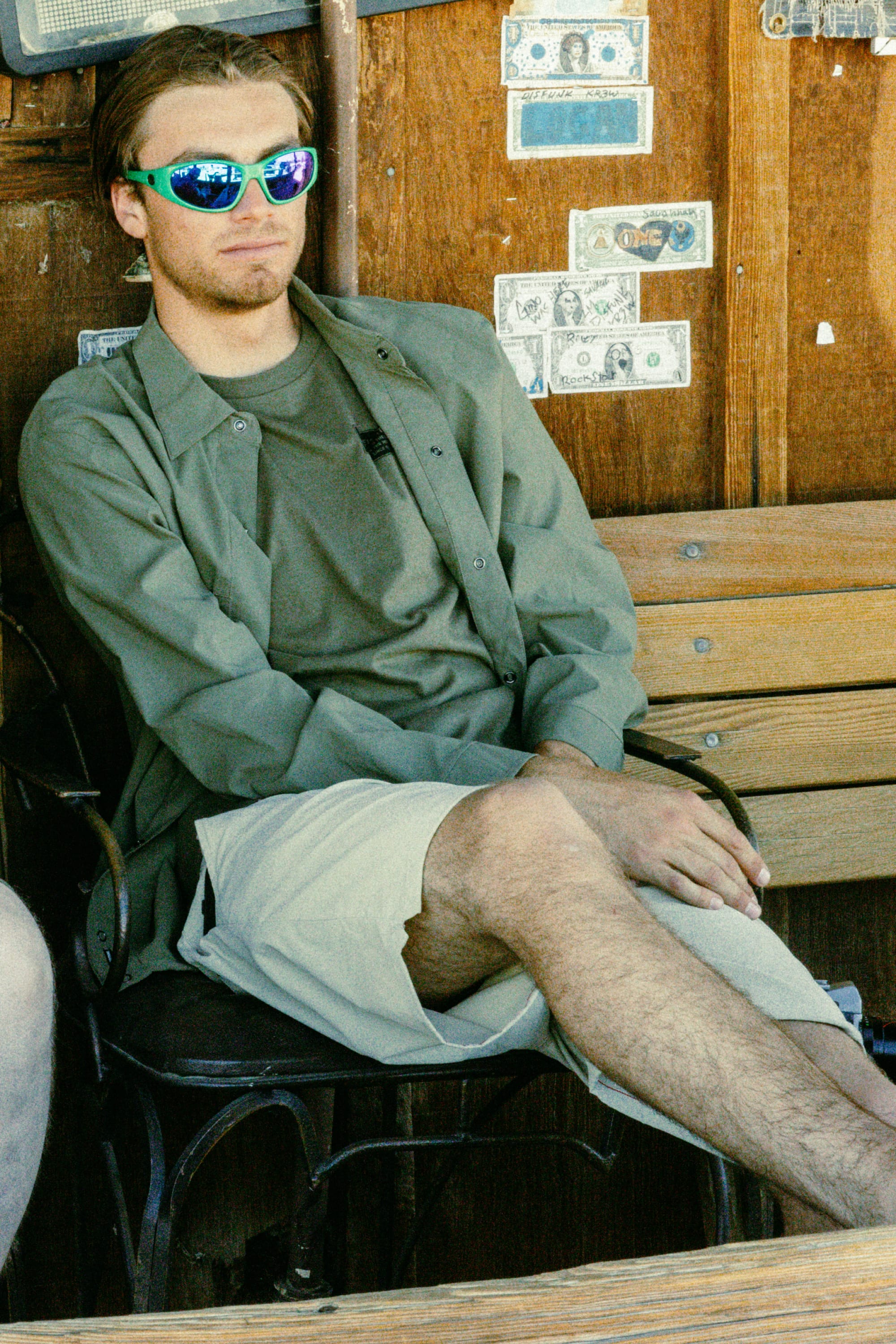 A young man wearing reflective sunglasses, a green shirt, and 686 Men's Everywhere® Featherlight Chino Shorts sits cross-legged on a wooden chair in front of a wall decorated with dollar bills.