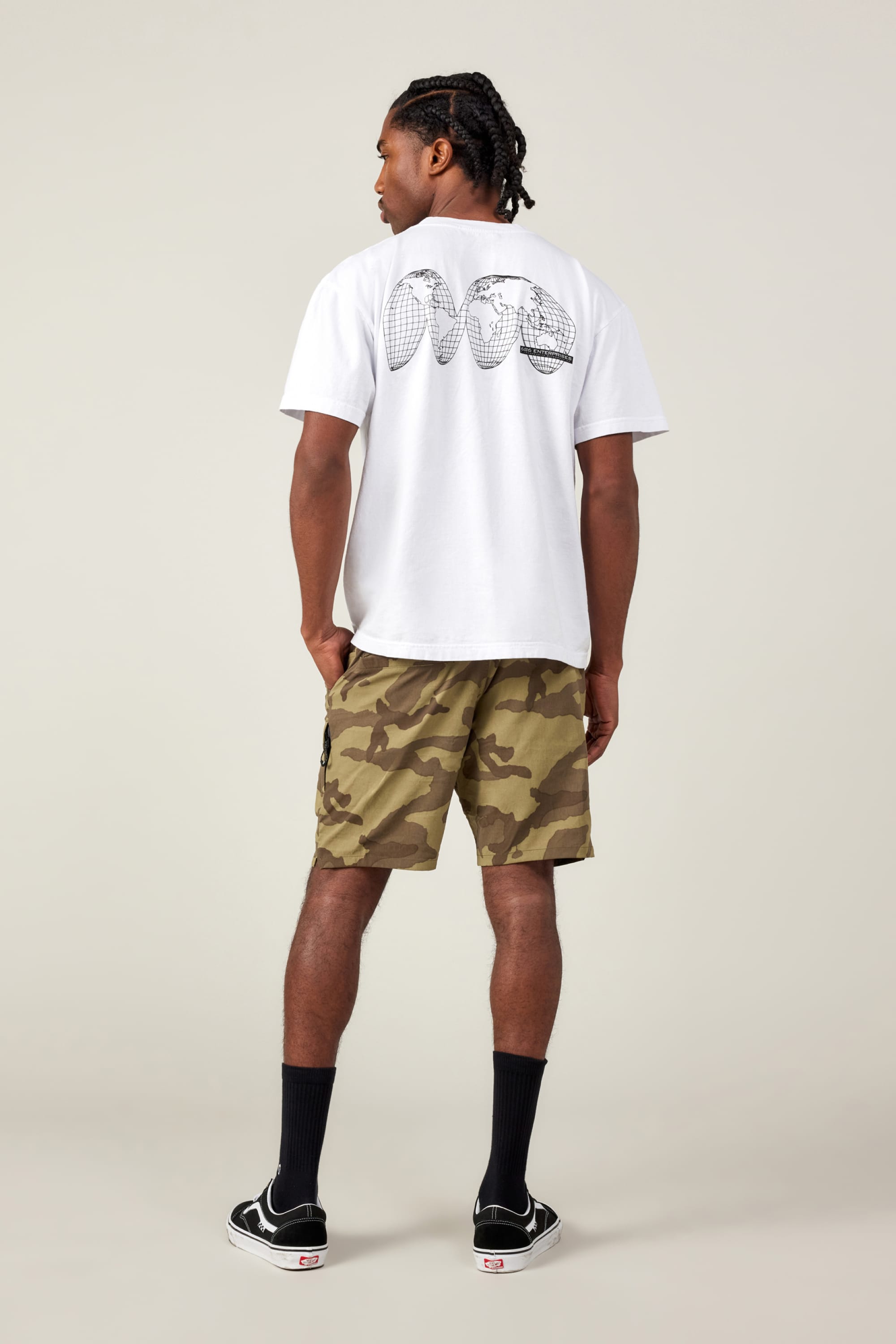 A man faces away, wearing a white tee with a back graphic, 686 Men's Everywhere® Featherlight Chino Short in camo, black socks, and black Vans sneakers. The background is light and plain.