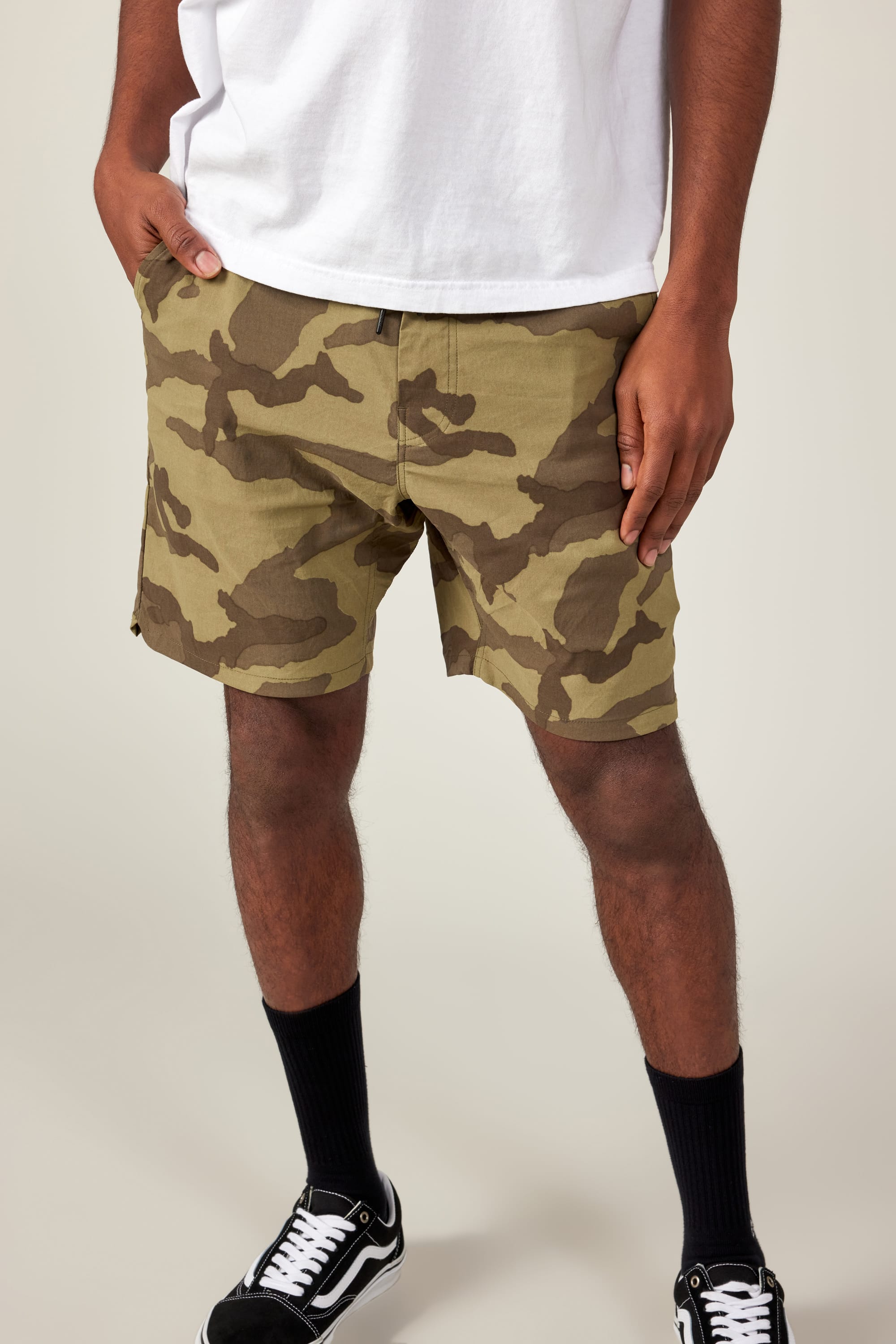 A person wearing a white T-shirt, 686 Men's Everywhere® Featherlight Chino Short by 686 in camouflage, black crew socks, and black-and-white Vans sneakers stands with one hand in their pocket against a plain background.