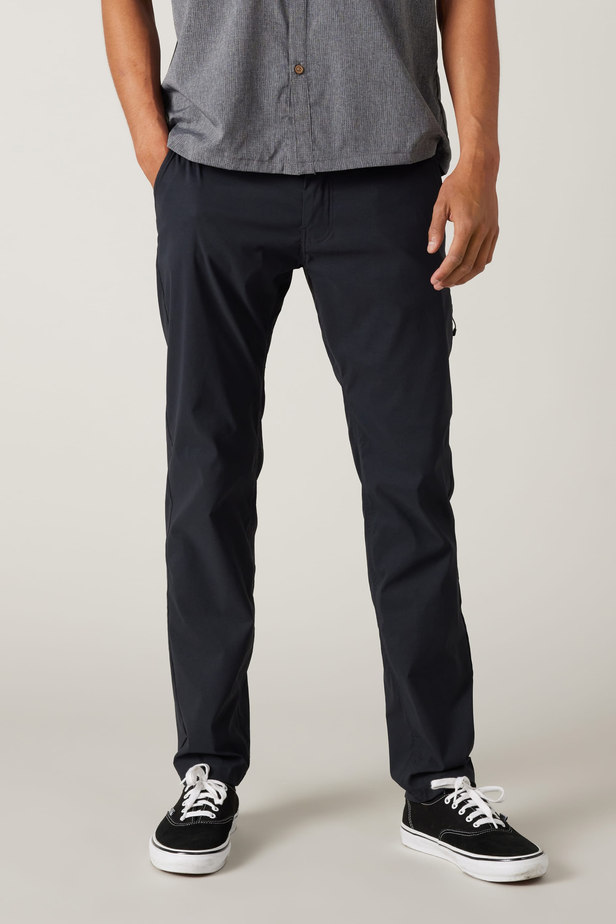 A person wears a gray short-sleeve button-up shirt, 686 Men's Everywhere® Featherlight Chino Pant - Slim Fit in black, and black-and-white sneakers with hands in pockets—a relaxed, breathable look for an easy summer wardrobe.