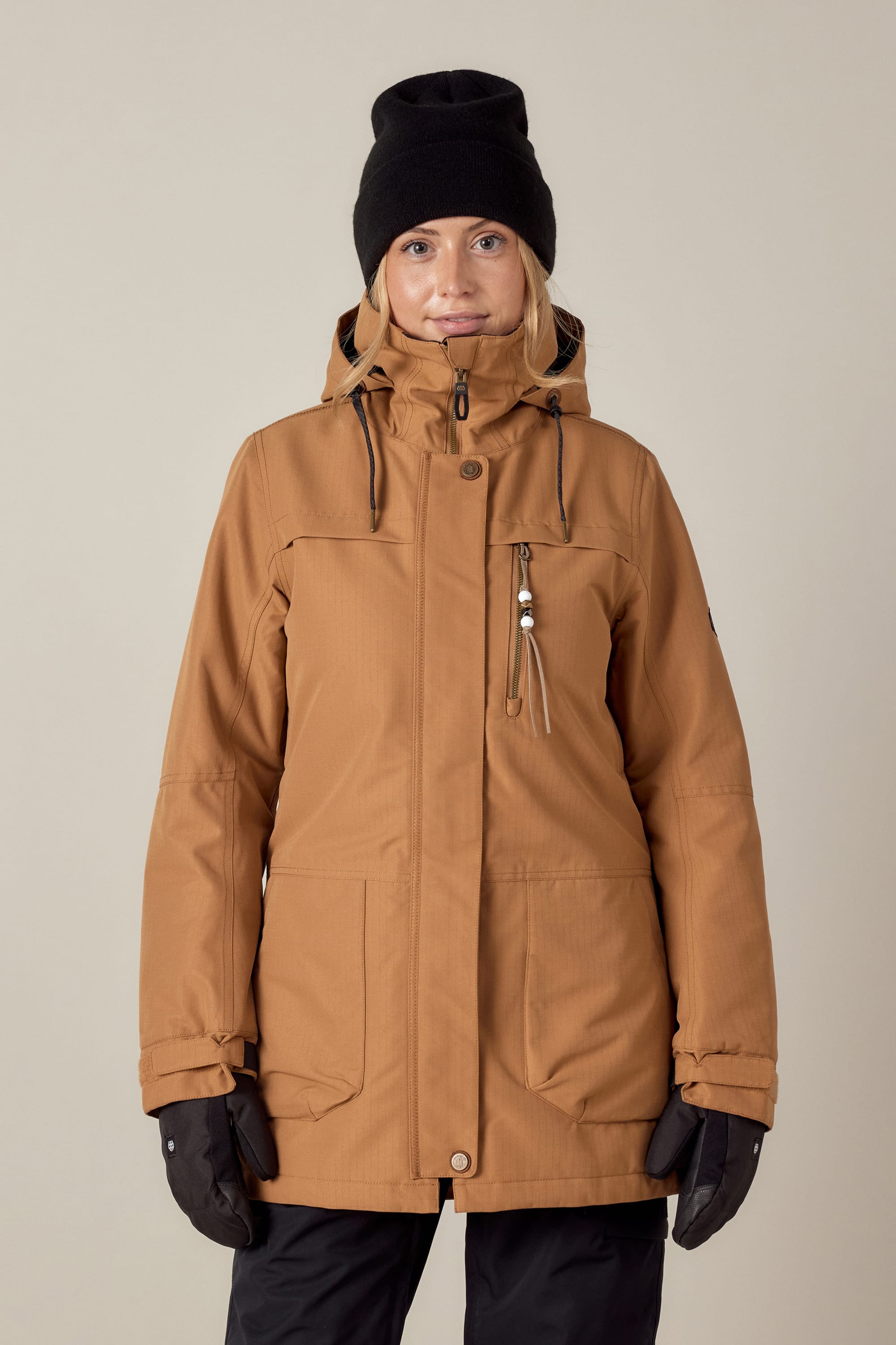 A person wearing the 686 Womens Spirit Insulated Jacket by 686, along with a black beanie and gloves, stands facing the camera against a plain light background.