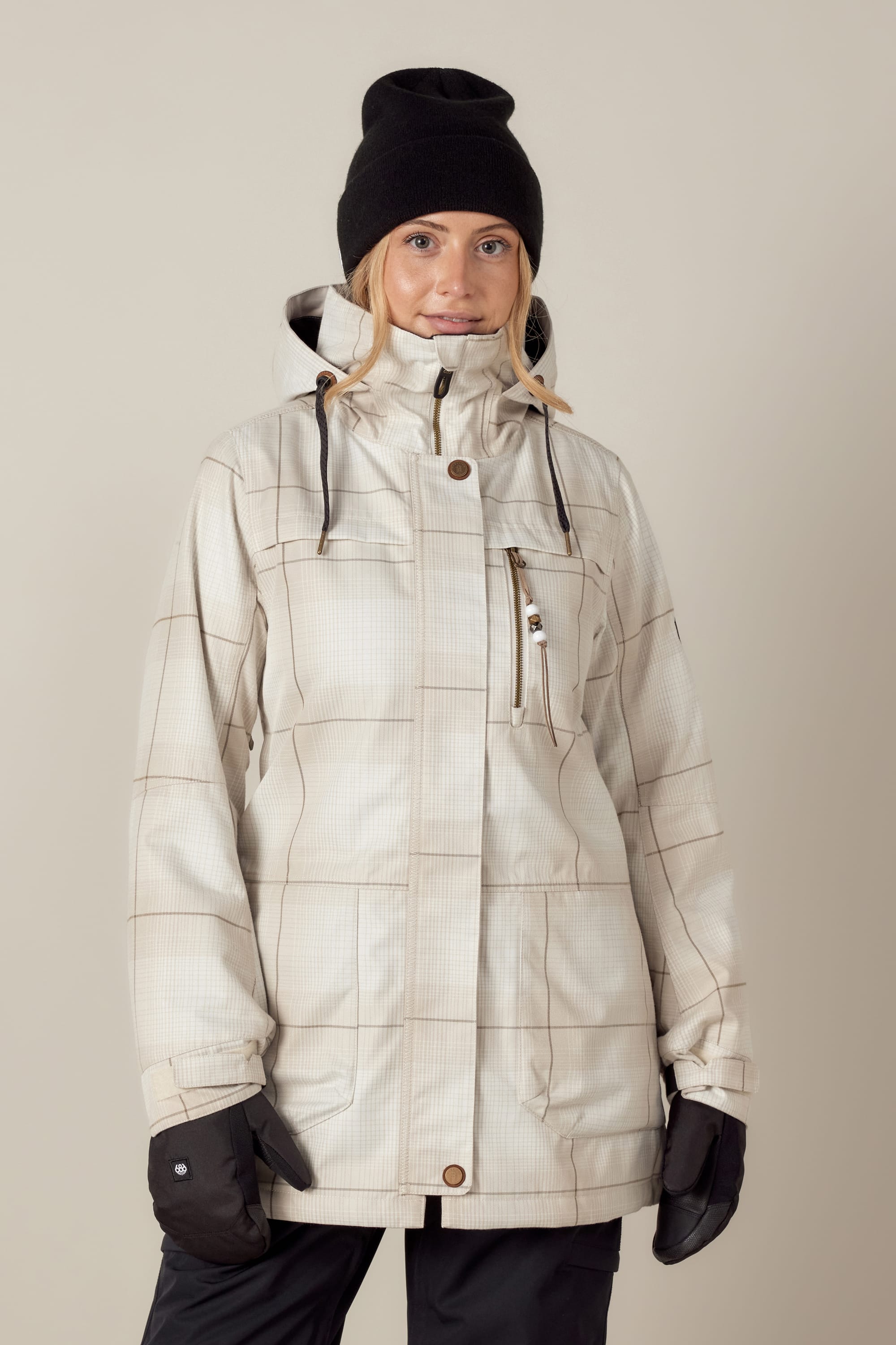 A woman wearing the 686 Womens Spirit Insulated Jacket by 686, featuring a light grid pattern and high collar, pairs it with black gloves and a beanie as she stands against a neutral background, facing forward with a slight smile.