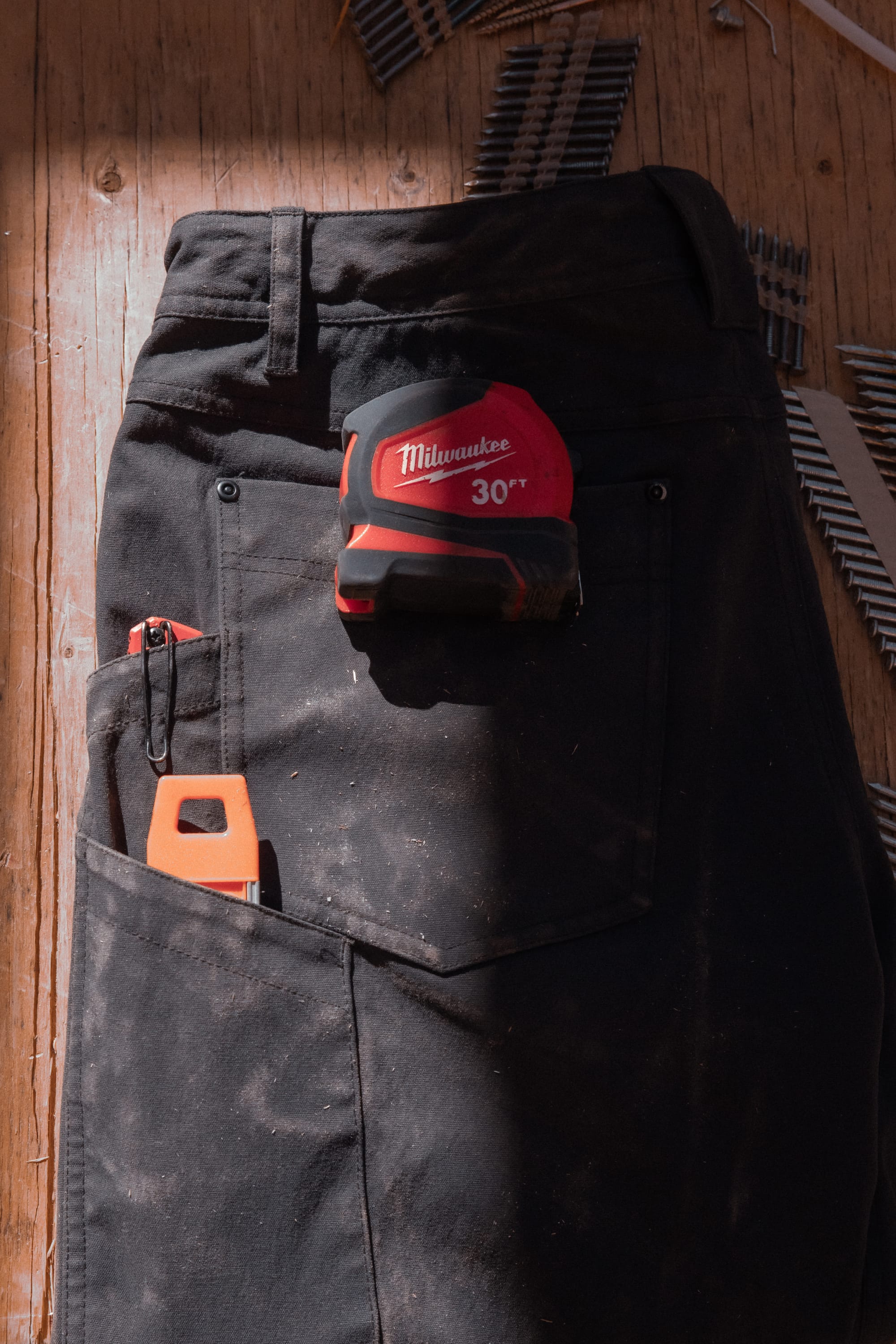 A pair of 686® Men's Unwork® Pant - Slim Fit by 686 lies dusted on a wooden floor, with a red Milwaukee tape measure and an orange utility knife in the pockets and metal nails scattered nearby.