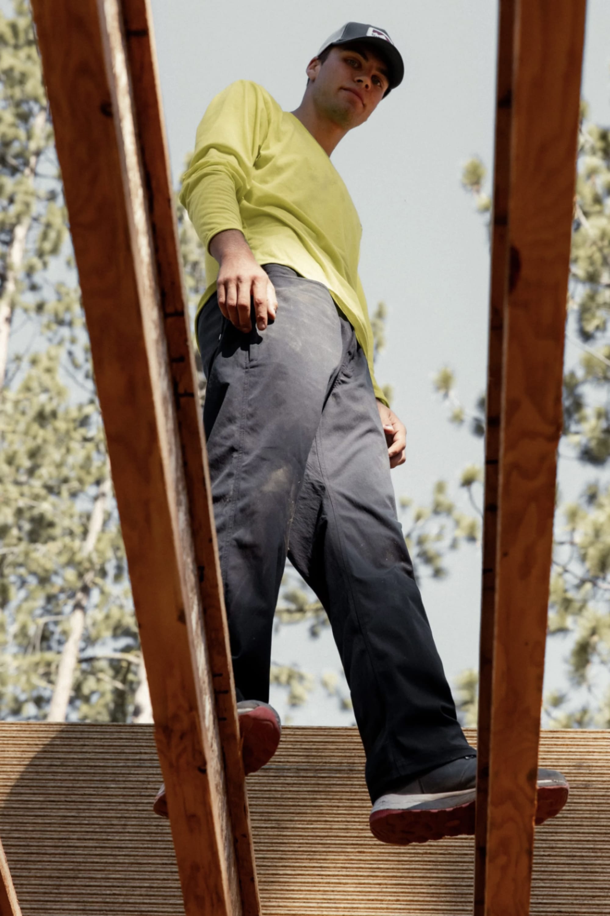 Wearing a yellow shirt, a classic workwear cap, and 686® Men's Unwork® Pant - Slim Fit, a person stands on wooden beams at a construction site, viewed from below with trees and sky in the background.