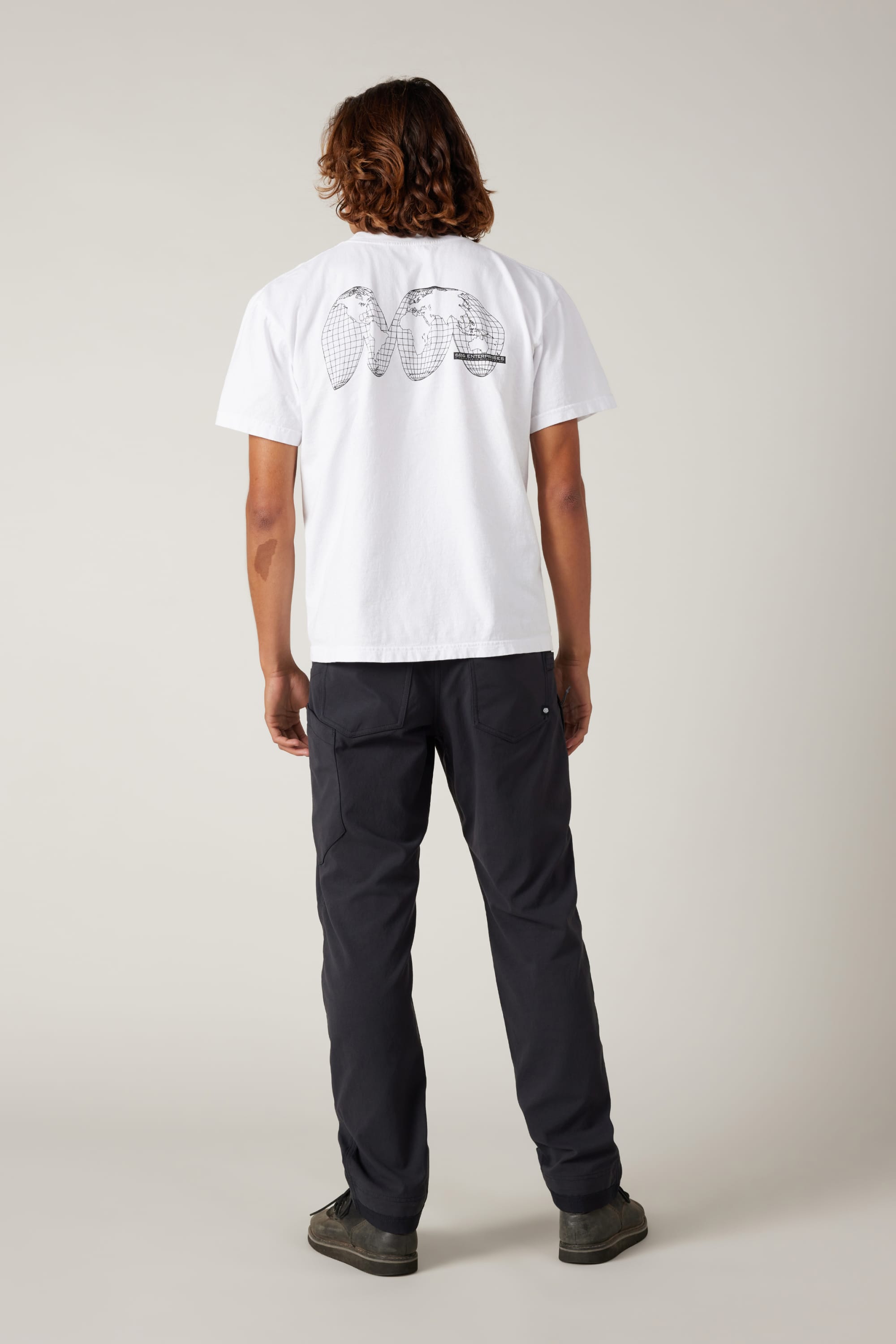 A person with shoulder-length hair faces away, wearing a white tee with black tennis racket graphics and 686® Men's Unwork® Pant - Slim Fit by 686, against a plain light background.