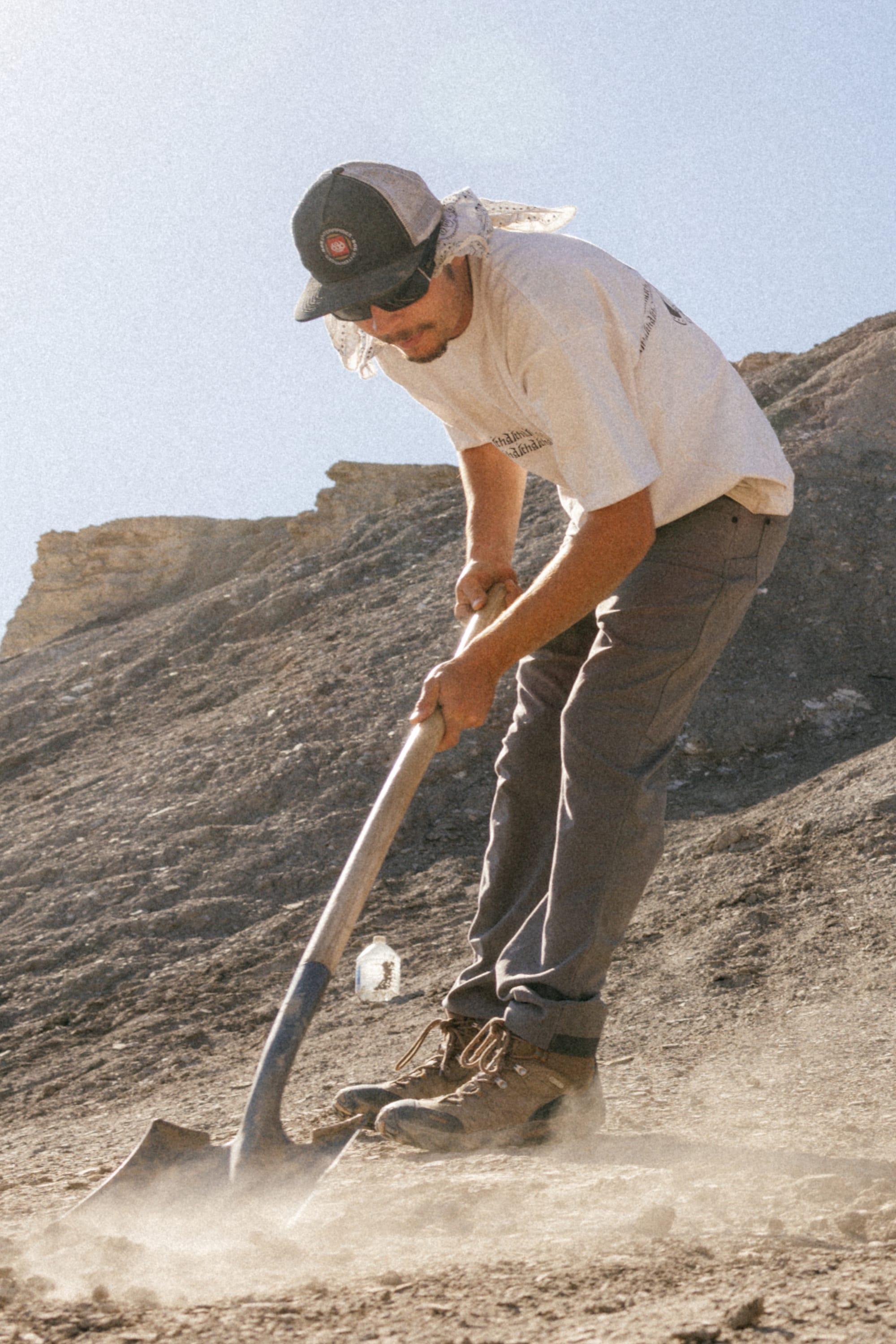 Wearing a cap, sunglasses, bandana, t-shirt, boots, and 686® Men's Unwork® Pant - Slim Fit by 686, a person uses a shovel to dig into dry, rocky ground on a hillside beneath bright sunlight.