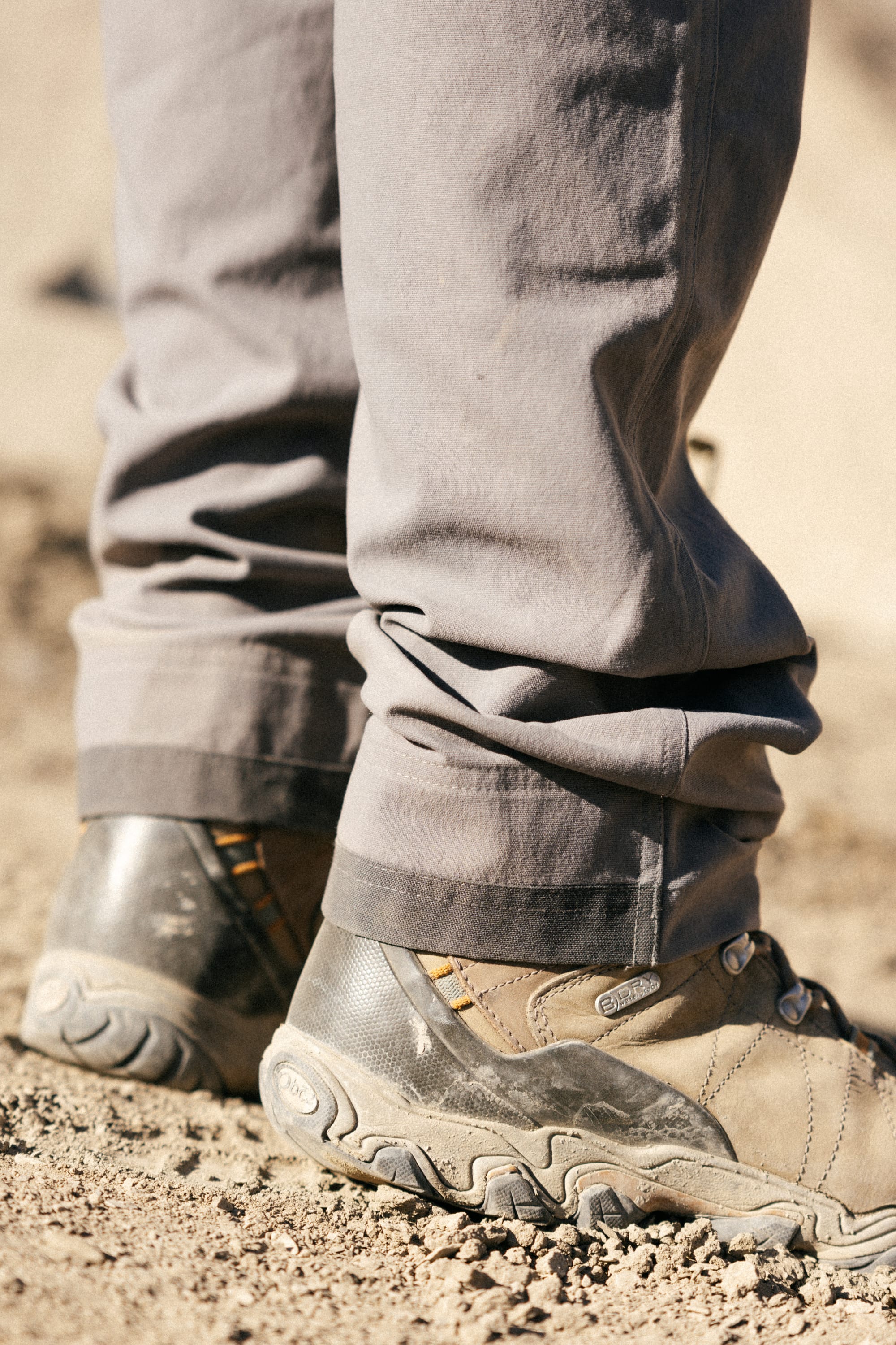 A close-up of a person wearing the 686® Men's Unwork® Pant - Slim Fit by 686 with beige hiking boots on sandy, dusty ground. The slightly crumpled pant legs partially cover the sturdy boots.