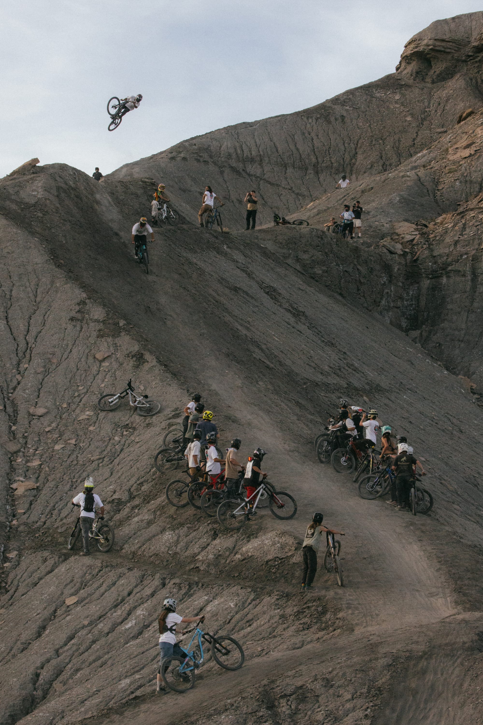 A group of mountain bikers on a rocky hillside watch a rider perform a high jump while others, dressed in 686® Men's Unwork® Pant - Slim Fit, wait or ride up the dusty trail below.
