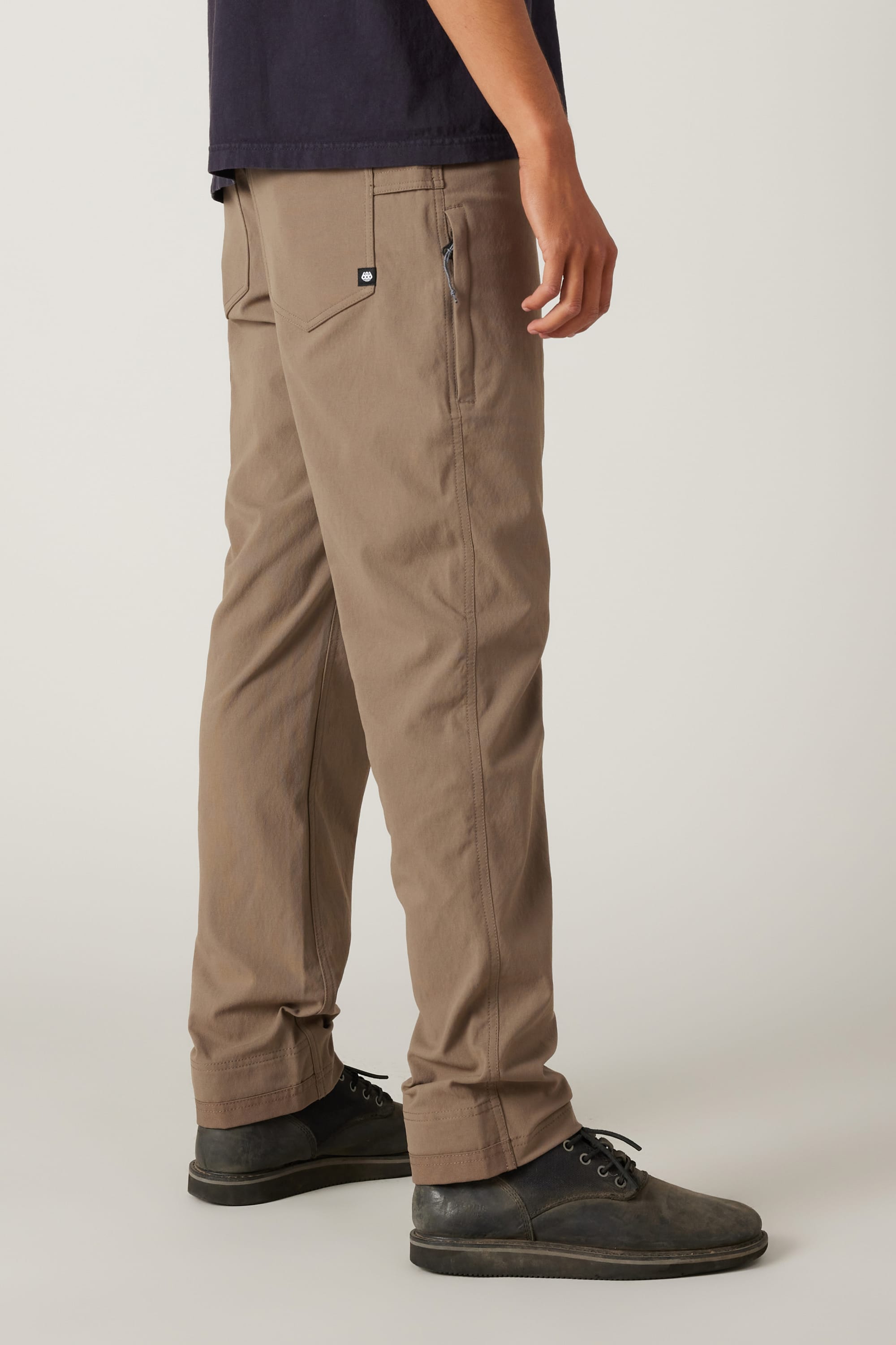 A person wearing 686® Men's Unwork® Pant - Slim Fit in light brown, paired with a dark navy shirt and black shoes, stands against a plain light background. Only the lower half of the body and right arm are visible.