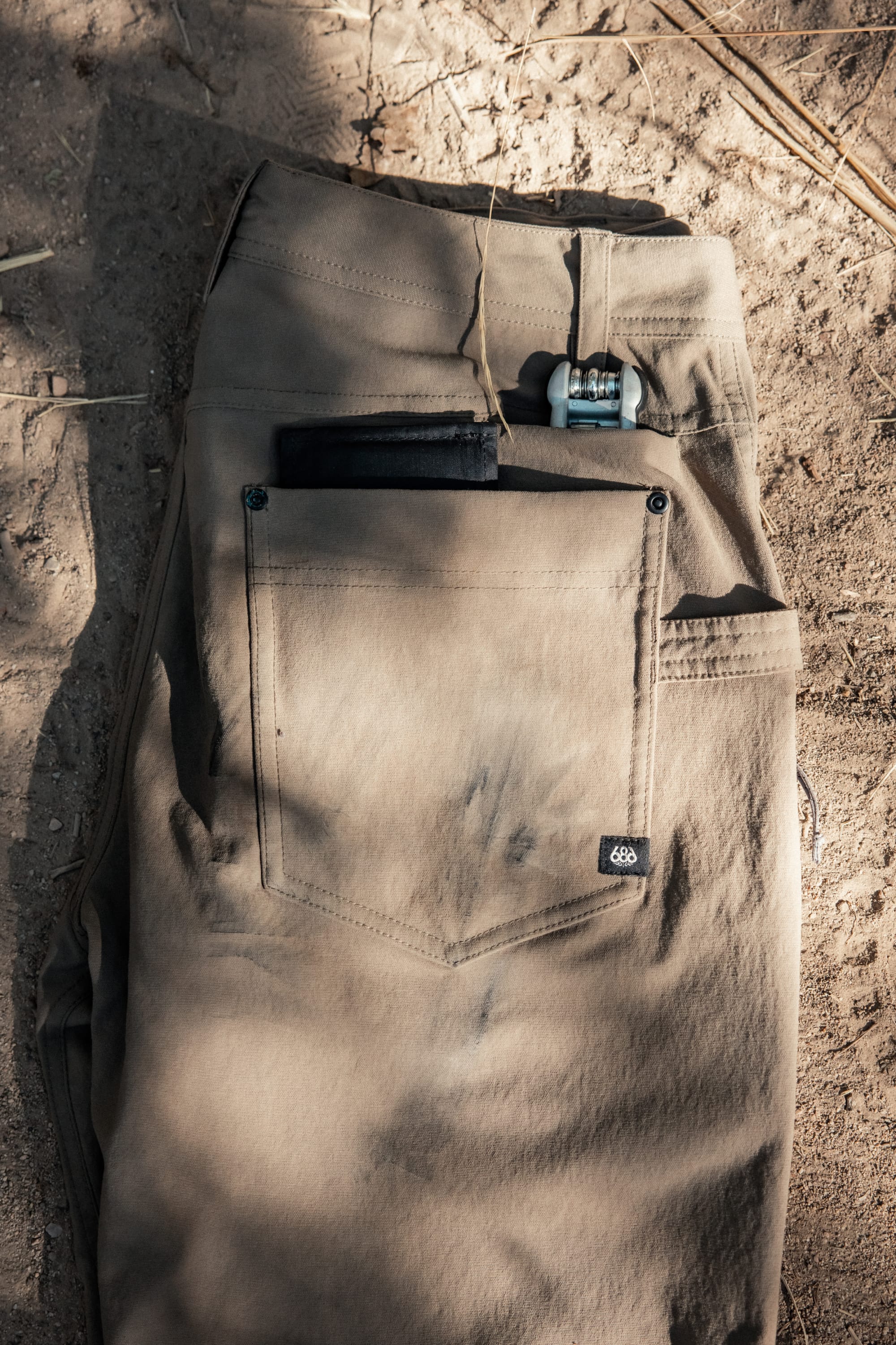 A pair of 686® Men's Unwork® Pant - Slim Fit in tan is folded on sandy ground. A black wallet and multi-tool peek from the back pocket, with sunlight and shadows highlighting the durable fabric by 686.