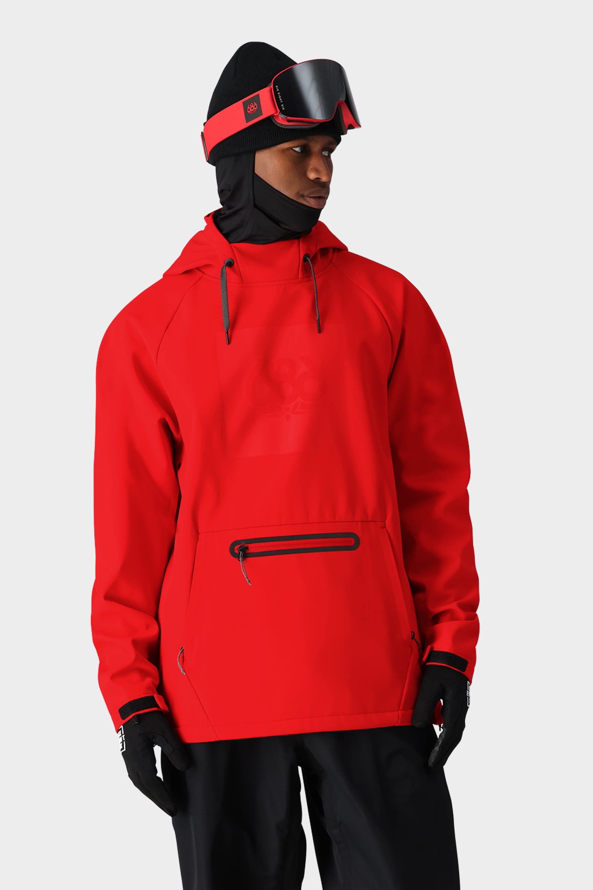 A person in a 686 Mens Waterproof Hoody stands against a plain light background, paired with black ski pants, gloves, a black balaclava, and red ski goggles on their forehead.