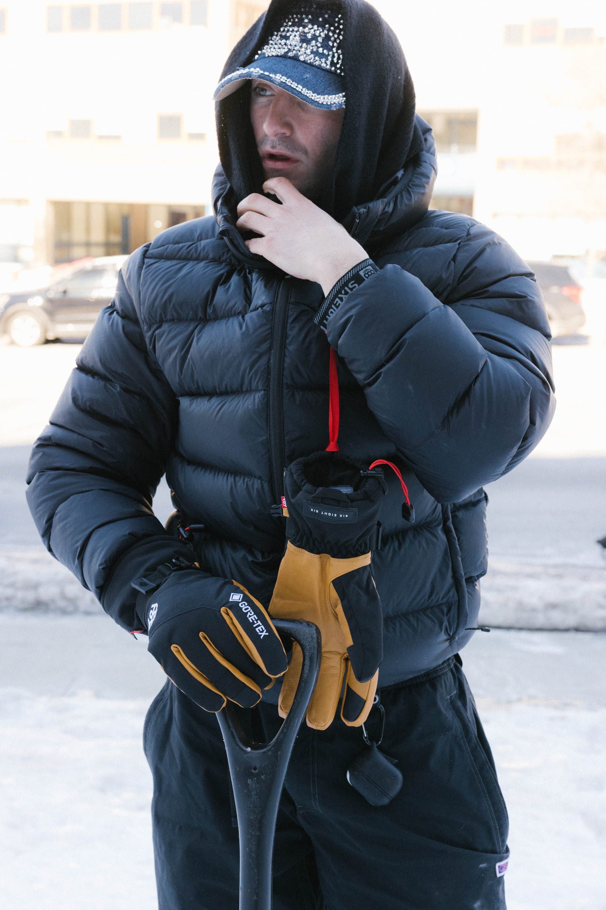 A person in a black puffer jacket and 686 Men's GORE-TEX Leather Linear™ Gloves stands on a snowy sidewalk, hood over cap, holding a shovel—prepared for outdoor work in cold weather.