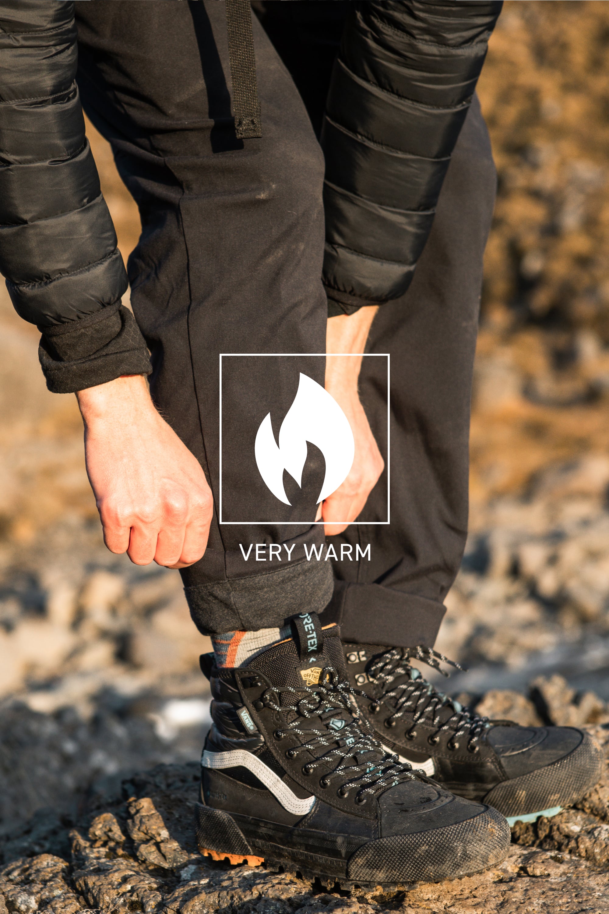 A person outdoors rolls up the cuff of 686 Men's Everywhere® Merino-Lined Pant - Slim Fit, staying warm and dry with a puffy jacket and rugged black boots. Text and a flame icon read VERY WARM, ideal for colder conditions. Rocky terrain appears in the background.