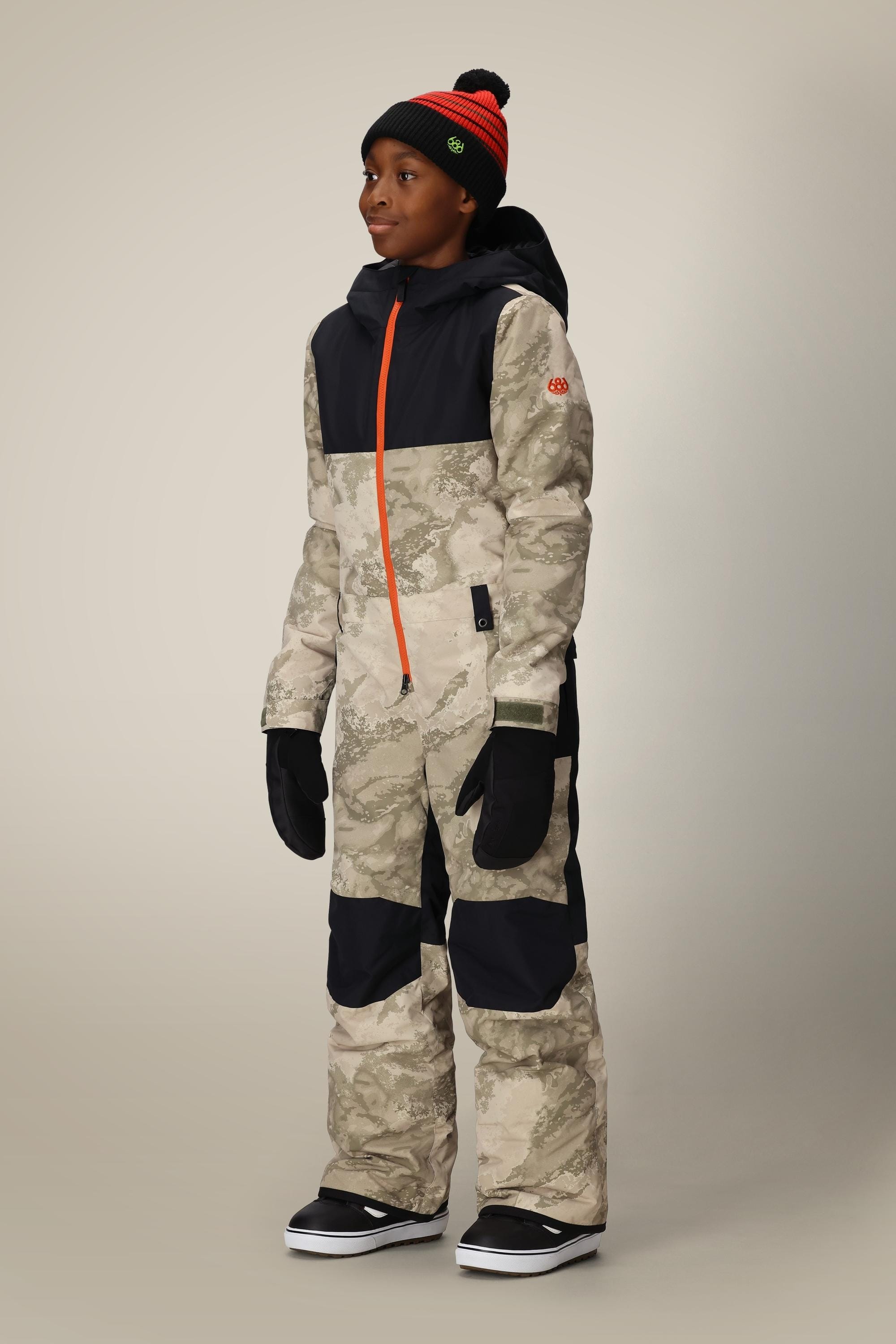 A young person wears the 686 Boys Shazam Insulated One-Piece—a camo-patterned polyester snowsuit with black panels, black hood and gloves, plus a black beanie with red stripe and pom-pom. The coverall features an orange front zipper.