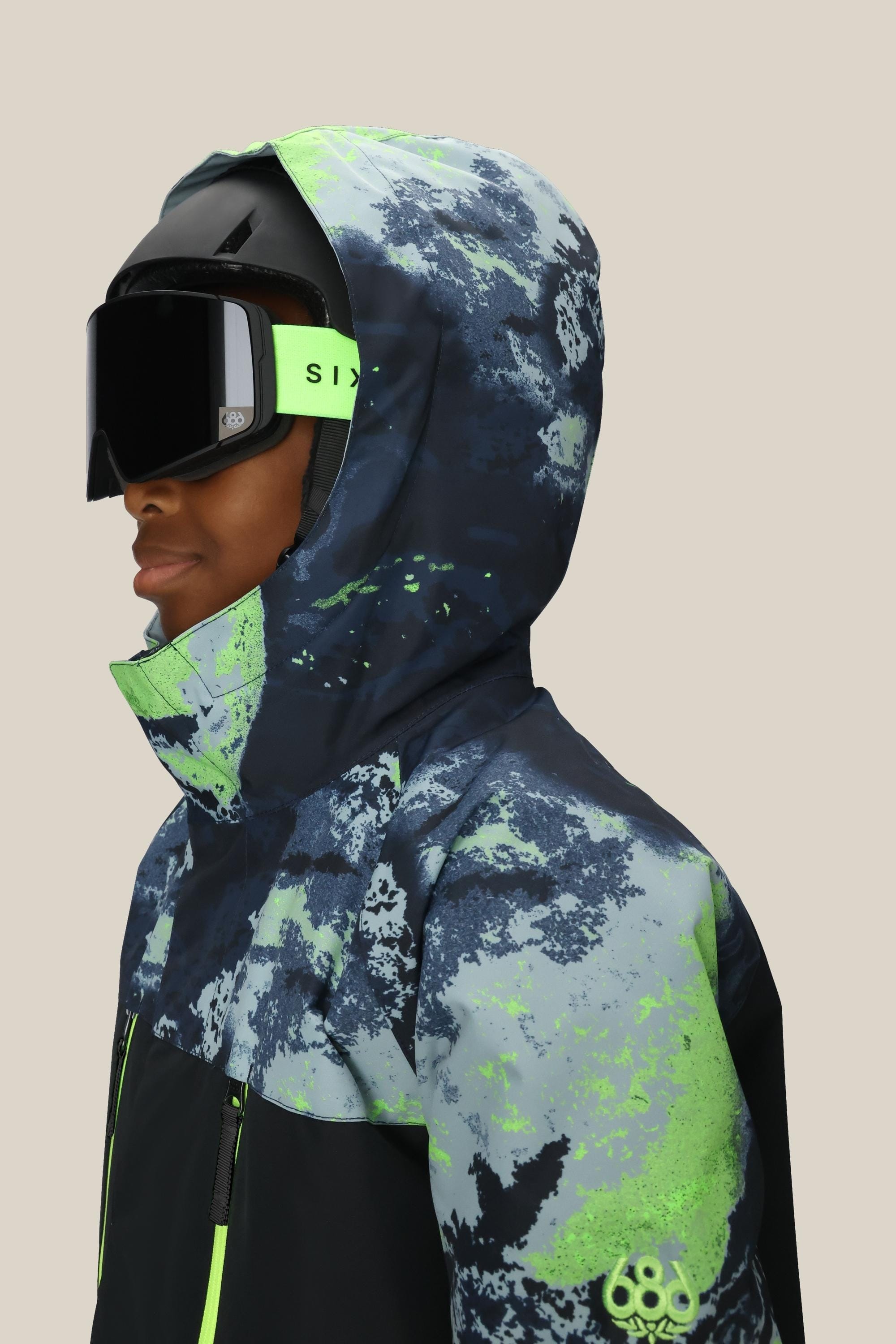 A boy is shown from the side in a 686 Boys' Geo™ Insulated Jacket, hood up, paired with black ski goggles and a black helmet. The blue and neon green geometric jacket by 686 highlights a tech-ready style against a plain background.