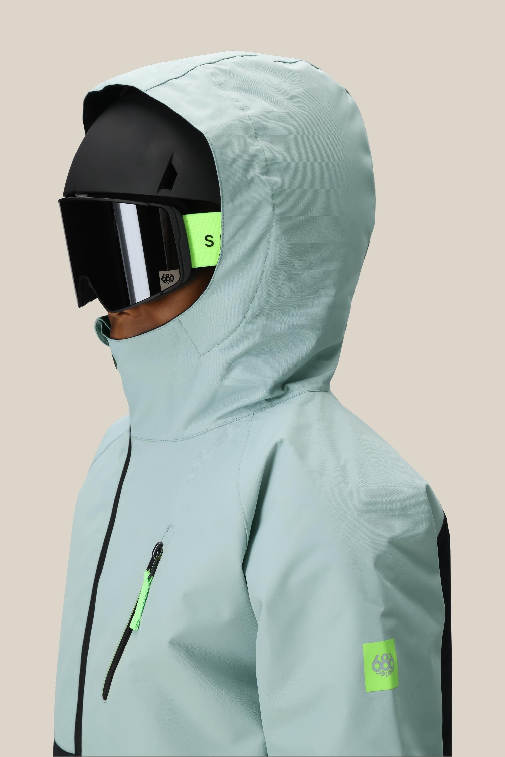 A boy wears the 686 Boys' Hydra™ Insulated Jacket by 686 in light blue with neon green accents, body-mapped lining, plus a black ski helmet and large goggles, facing left against a plain beige background.