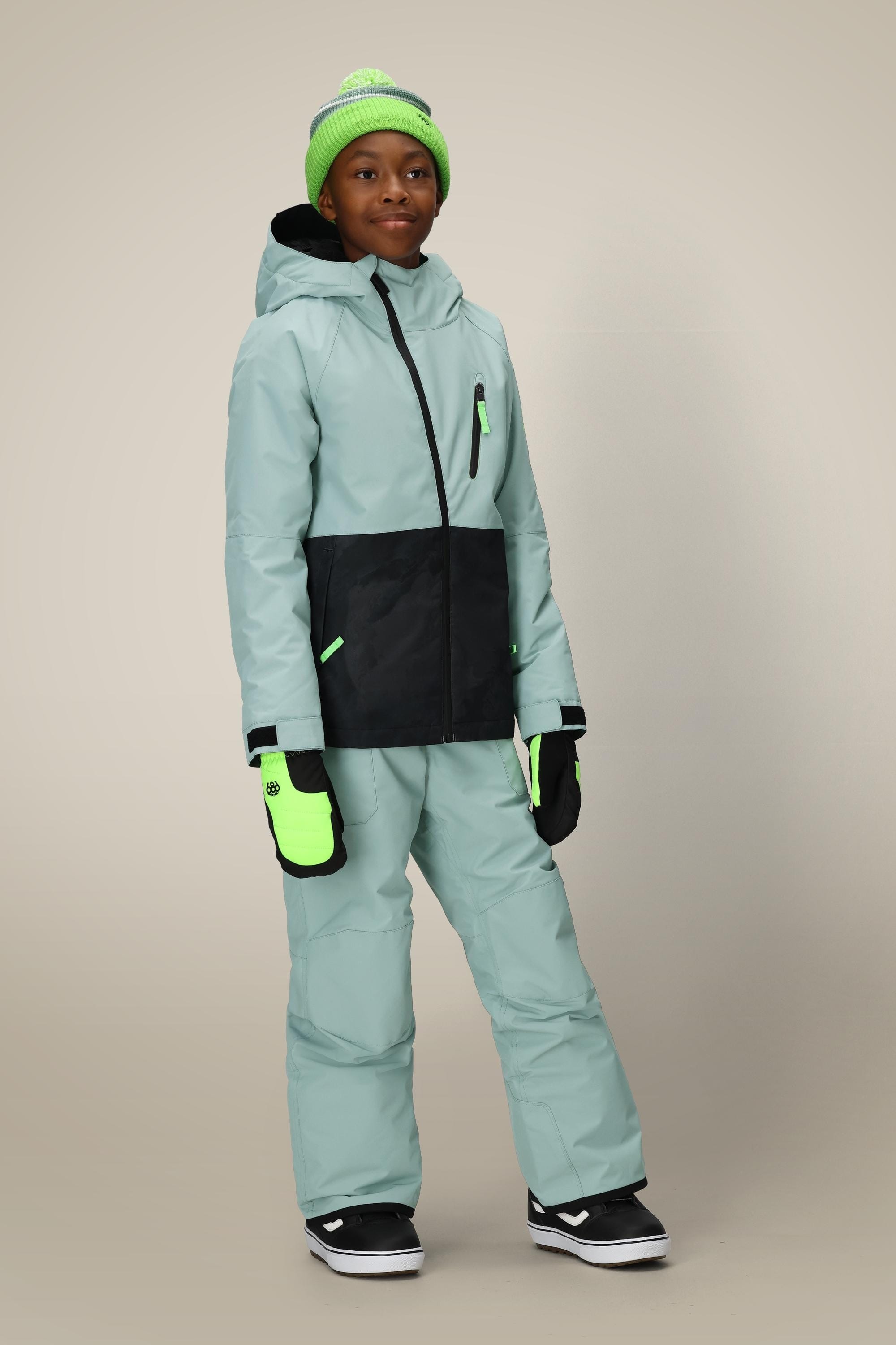 A child in the 686 Boys' Hydra™ Insulated Jacket, coordinated blue and black pants, neon green beanie, and mittens stands against a plain beige background.