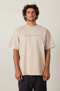 A man with curly brown hair wears the 686 Elements Relaxed S/S Tee in beige, featuring “SIX EIGHT SIX” on the front, paired with dark patterned shorts, standing against a plain light background.