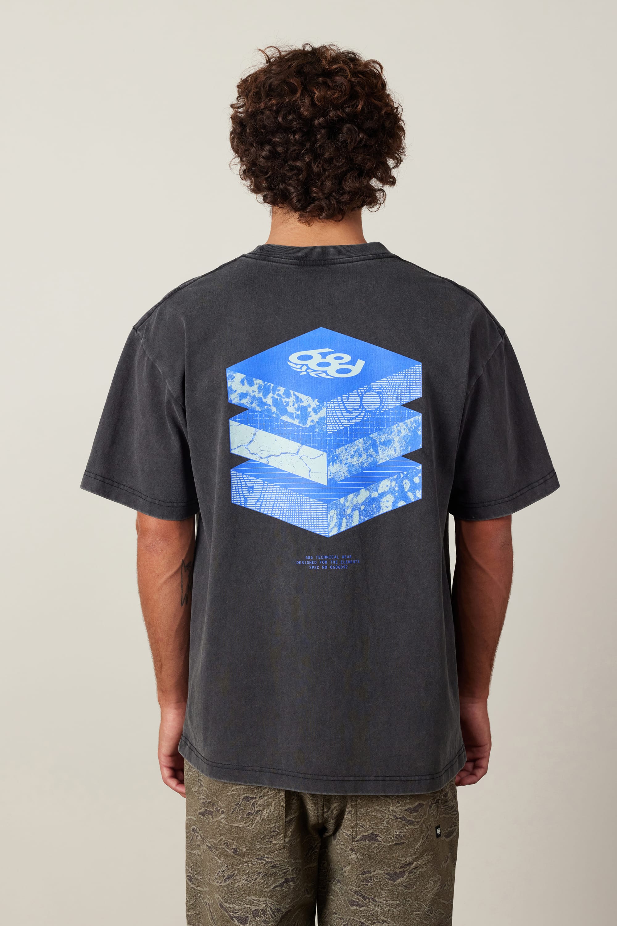 A person with curly hair is seen from behind, wearing the 686 Elements Relaxed S/S Tee by 686, featuring a blue and white geometric cube design on the back and paired with casual patterned pants.
