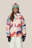A person smiles, wearing the 686 Girls' Athena™ Insulated Jacket by 686 with colorful abstract patterns, a light blue beanie with a pink pom-pom, white snow pants, and black gloves against a plain beige background.