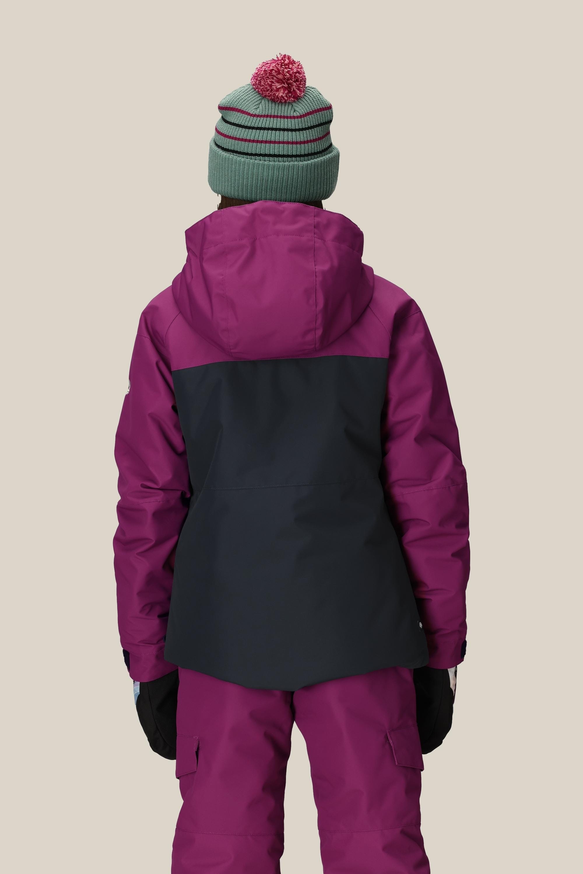 A person stands facing away, wearing the 686 Girls' Athena™ Insulated Jacket in purple and black by 686, with matching purple snow pants, black gloves, and a green beanie with a red pom-pom against a beige background.