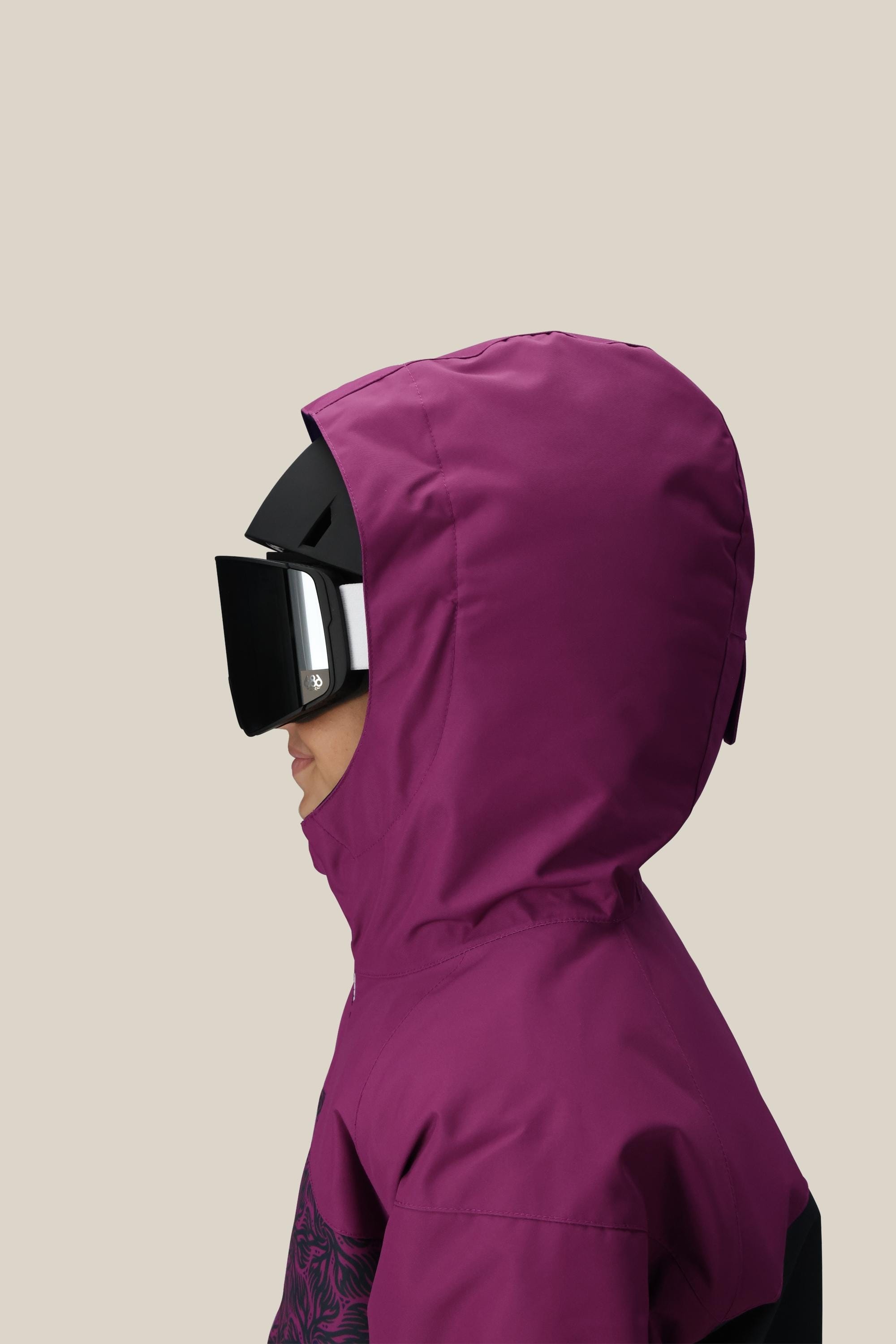 A girl in profile wears the 686 Girls' Athena™ Insulated Jacket in magenta with a hood and large black ski goggles, set against a plain beige background.