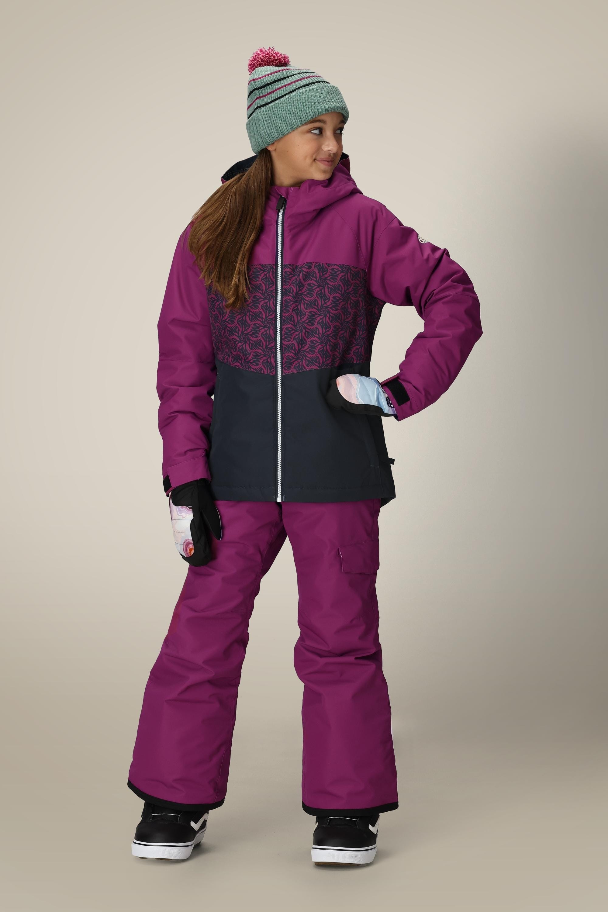 A girl stands smiling with one hand on her hip, wearing the 686 Girls' Athena™ Insulated Jacket in magenta and black, a teal knit hat, matching magenta snow pants, white gloves, and black snow boots.