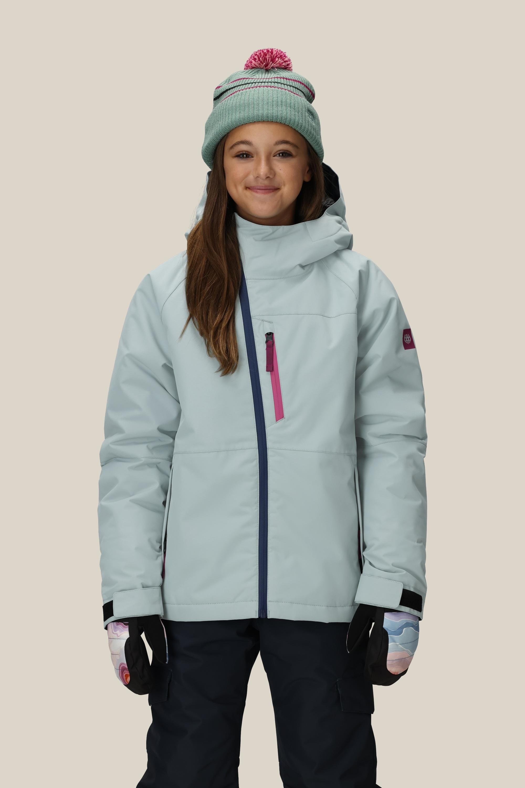 A smiling young girl wears the 686 Girls' Hydra™ Insulated Jacket by 686, paired with a mint green knit hat with a pink pom-pom, dark pants, and patterned gloves, standing against a plain beige background.