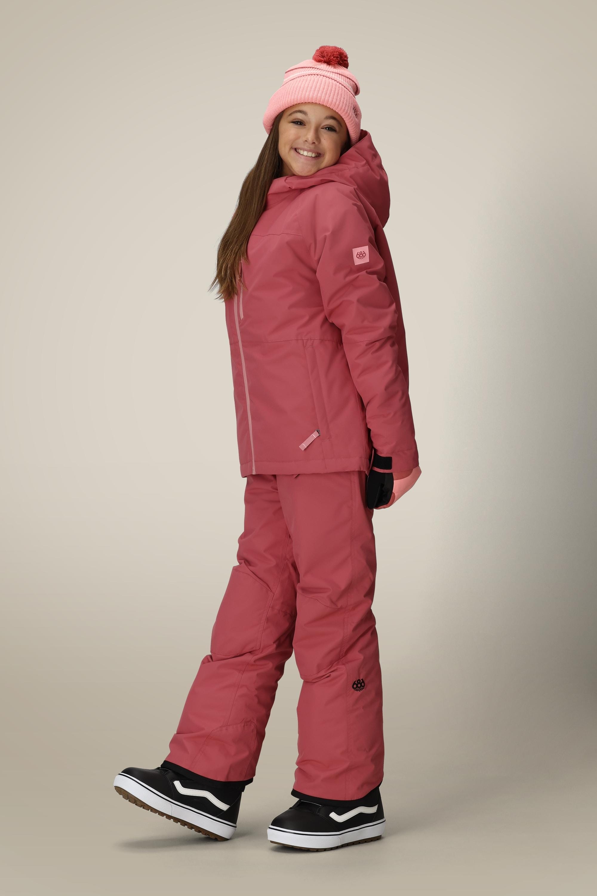 A smiling girl poses in the 686 Girls' Hydra™ Insulated Jacket, matching pink snow pants and beanie with pom-pom, black gloves, and black snow boots against a plain background.