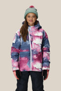 A girl smiles against a beige background in the 686 Girls' Hydra™ Insulated Jacket by 686, featuring pink, white, and blue patterns with body-mapped lining, black pants, pink gloves, and a light blue beanie topped with a pink pom-pom.
