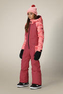 A young person in the 686 Girls Shine Insulated One-Piece by 686, paired with a pink beanie, black gloves, and black snow boots, stands slightly turned to the side against a plain beige background.