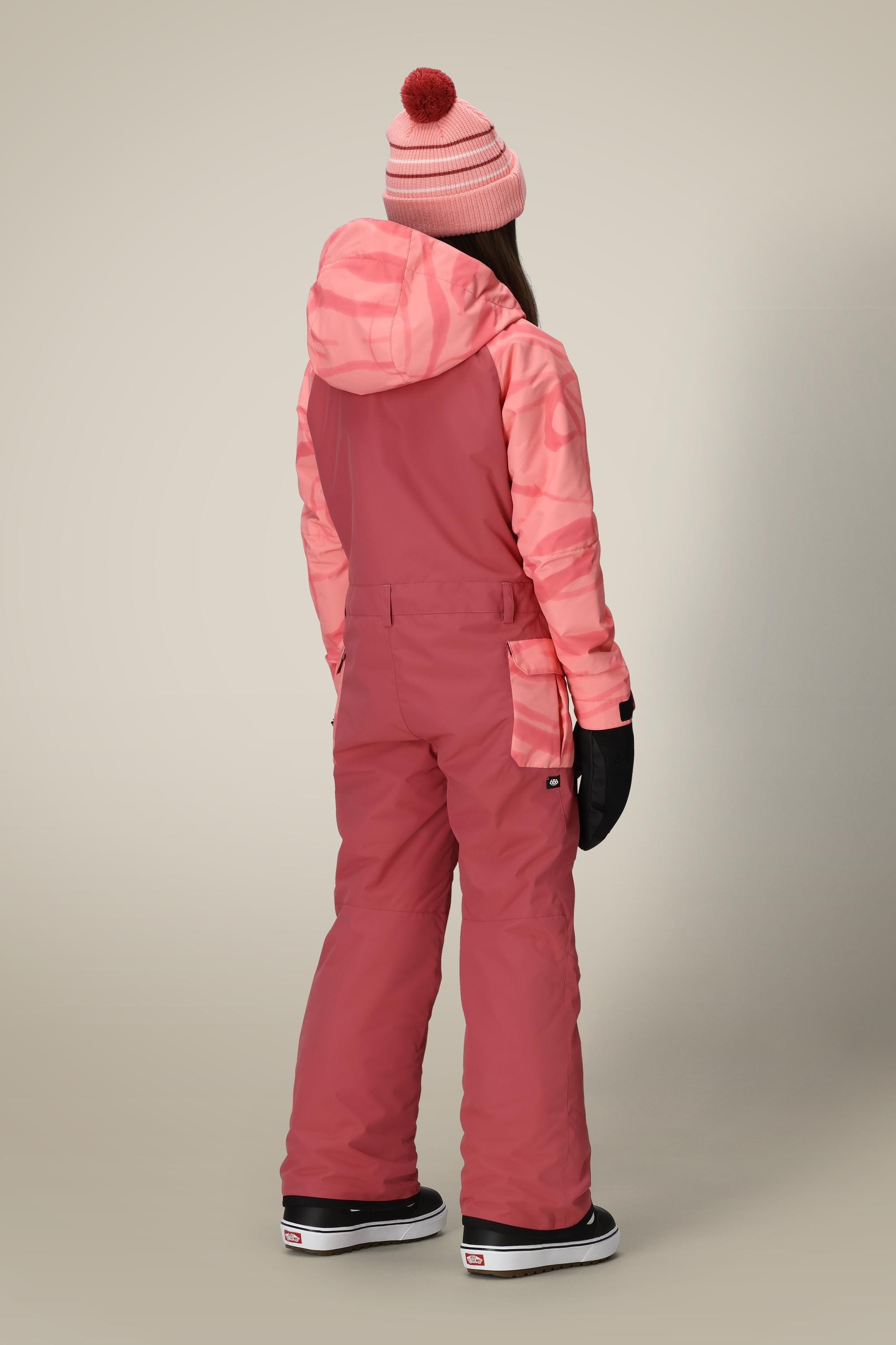 A person stands facing away in the 686 Girls Shine Insulated One-Piece, a pink and coral waterproof coverall from 686 with a patterned hood, black gloves, black boots, and a pink beanie with red pom-pom, on a beige background.