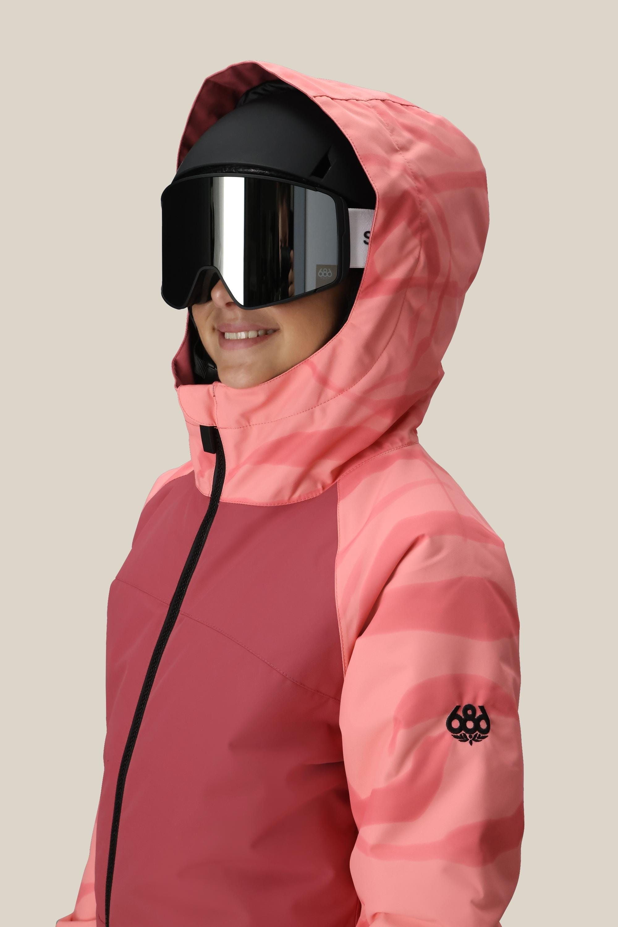 Wearing the 686 Girls Shine Insulated One-Piece and black ski goggles, a person in a pink hooded jacket smiles slightly to the side against a light backdrop, showcasing the cozy 686 style.