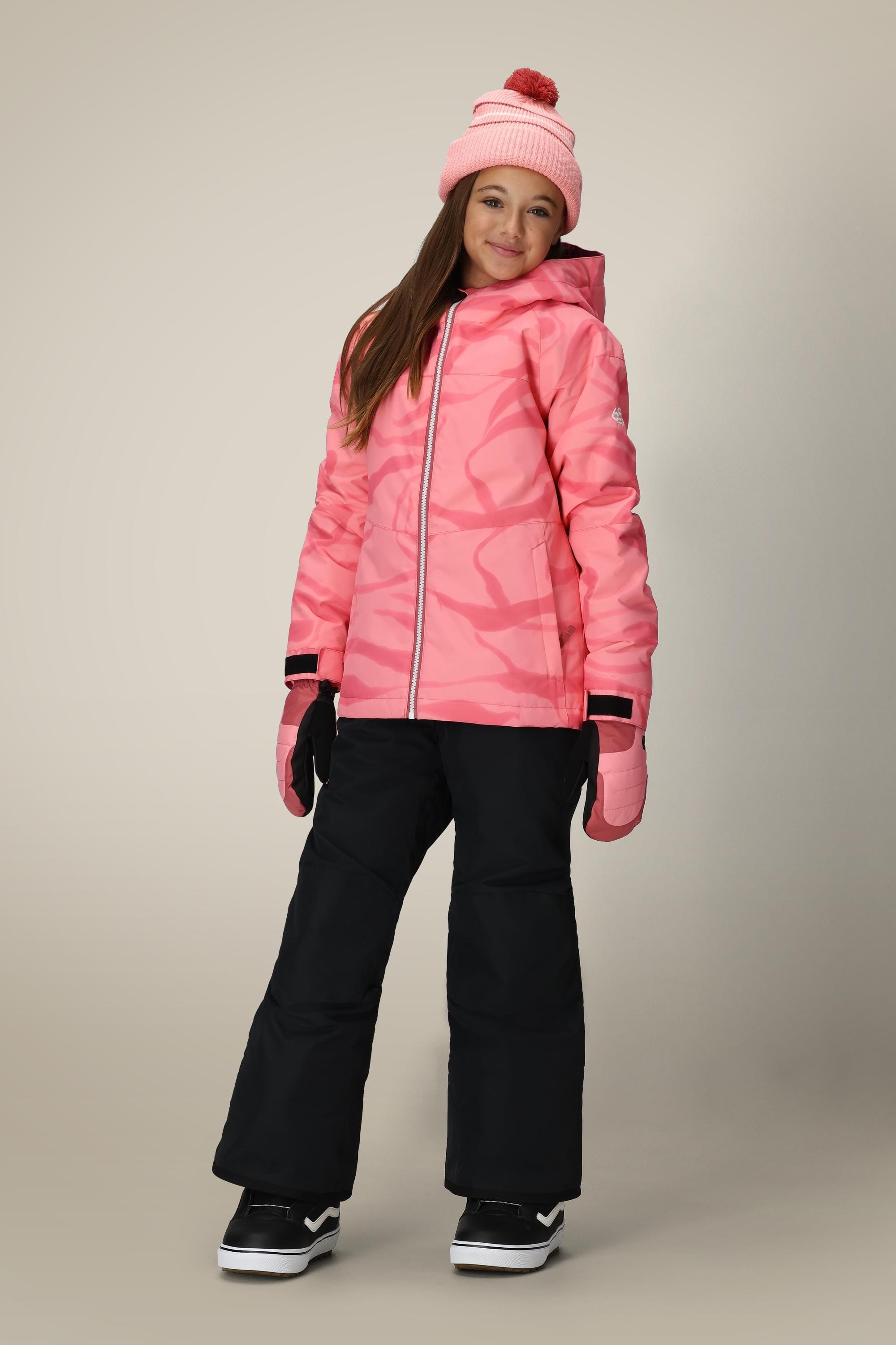 A young girl wears the 686 Girls Sierra Insulated Bib by 686, paired with a pink winter jacket, matching beanie, black gloves, and snow boots as she smiles at the camera against a plain background.
